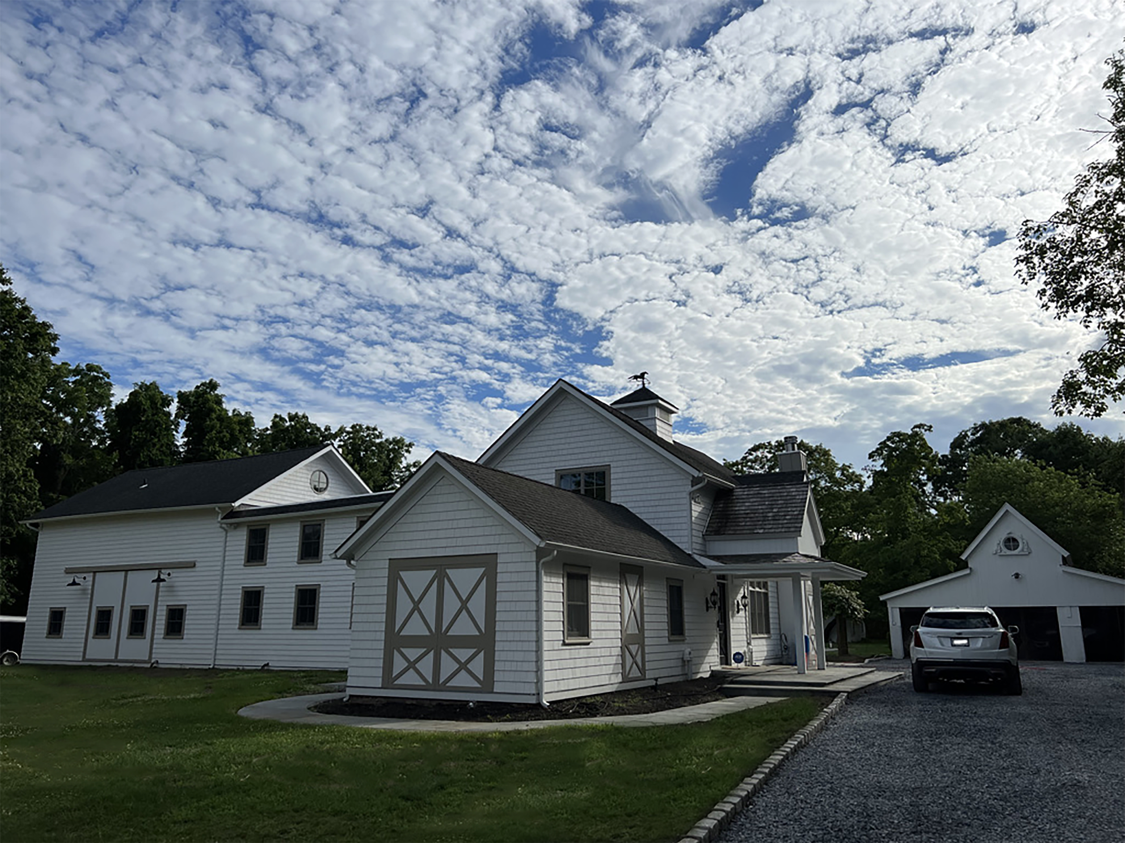 A white farmhouse with a barn and a gravel driveway sits under a cloudy sky.