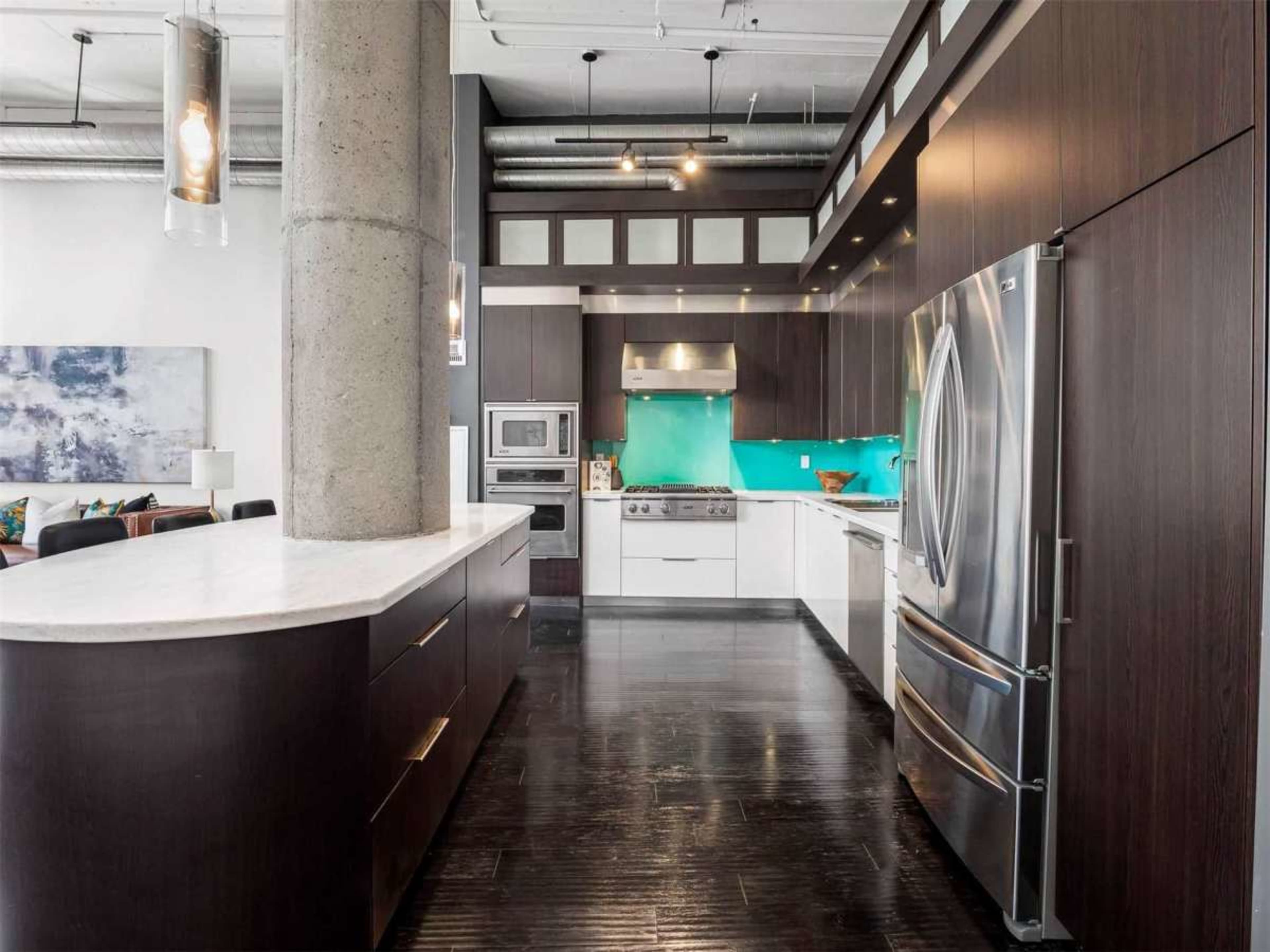 The image shows a modern kitchen with dark wood cabinetry, a central island, stainless steel appliances, and a bright turquoise backsplash.