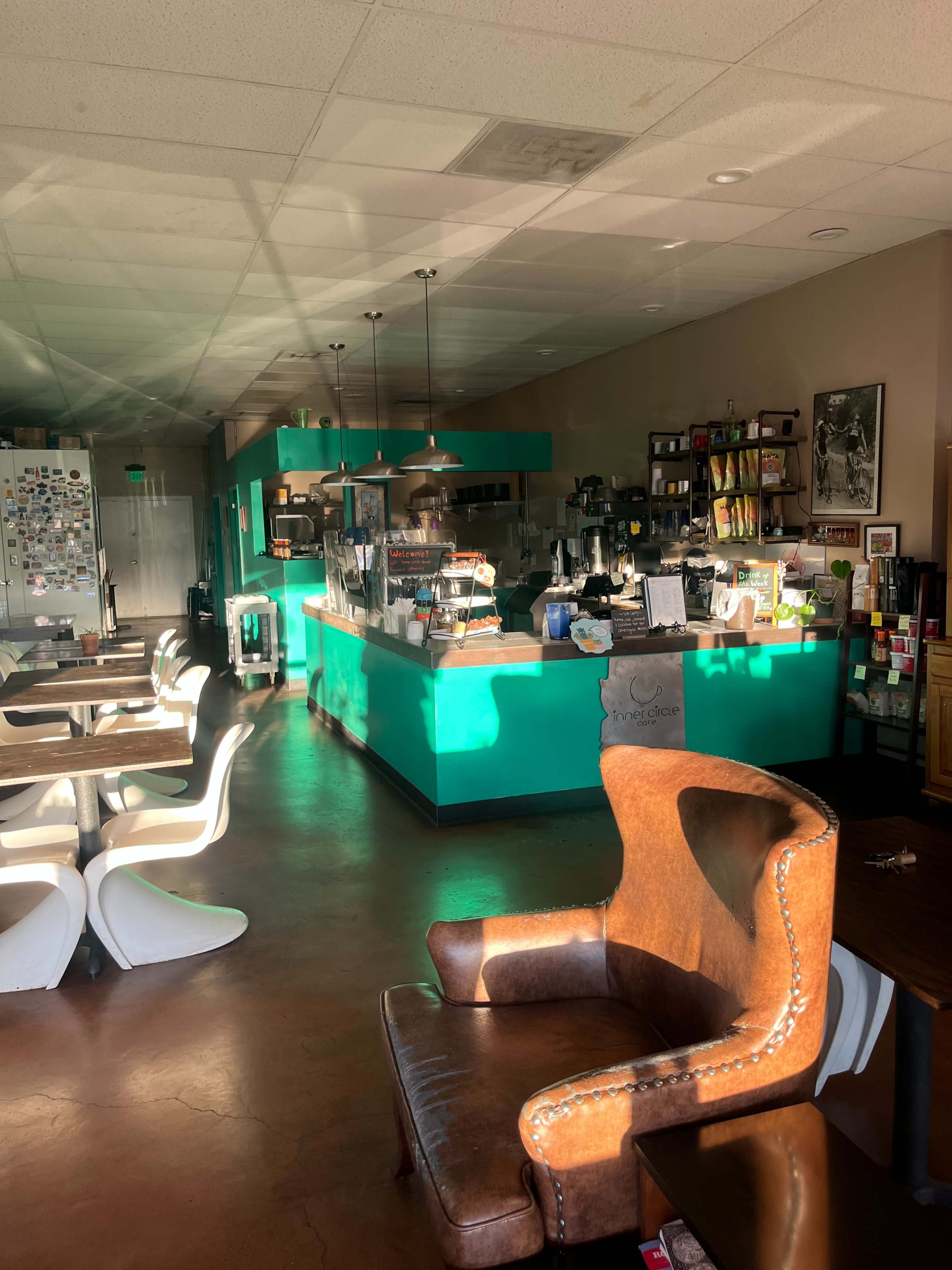 A cozy coffee shop with a turquoise counter, wooden tables, and white chairs, illuminated by natural light.