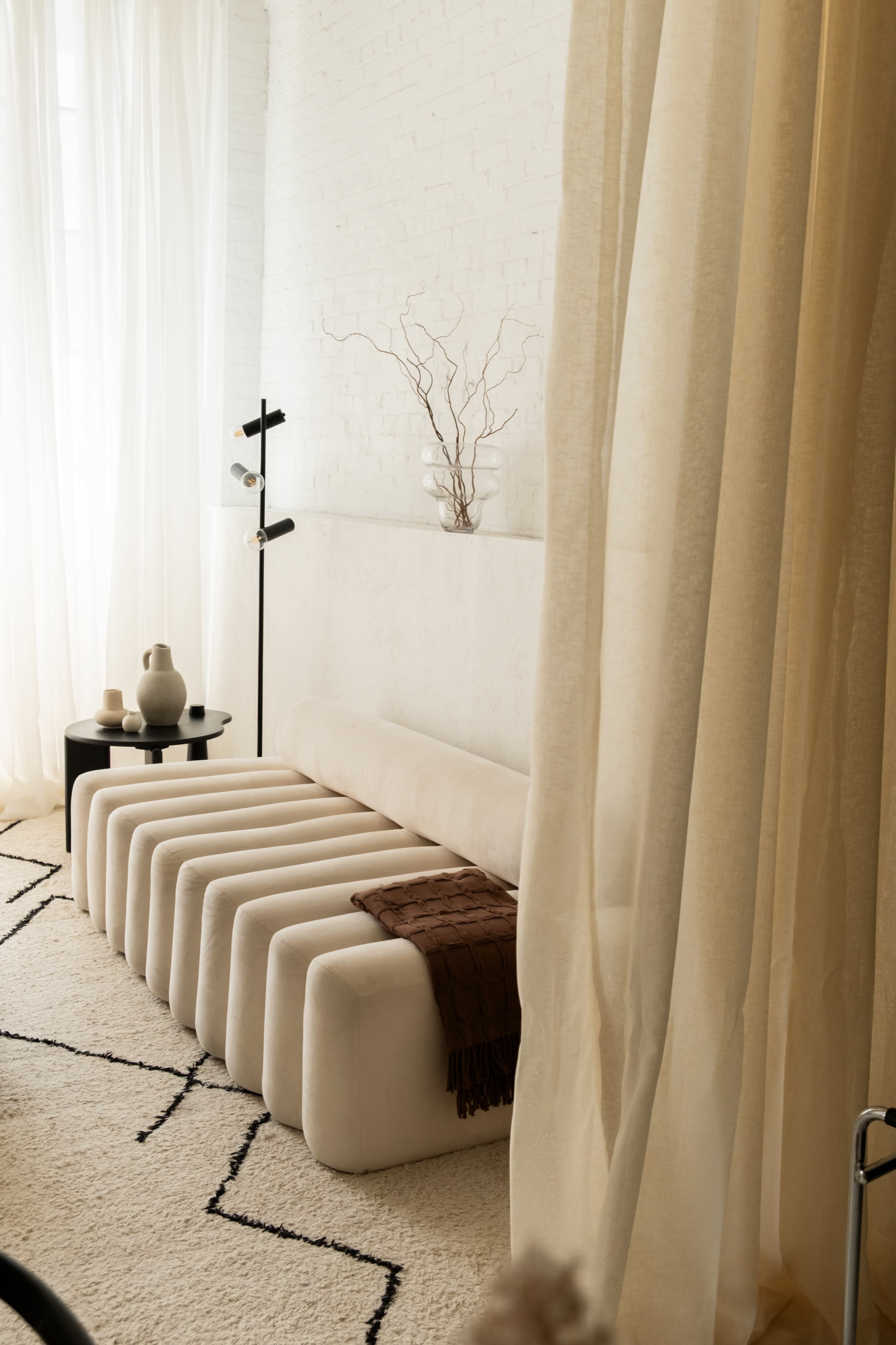 The image shows a minimalist living space featuring a long, padded sofa, a small black table with a vase, and sheer curtains that partially obscure the view.