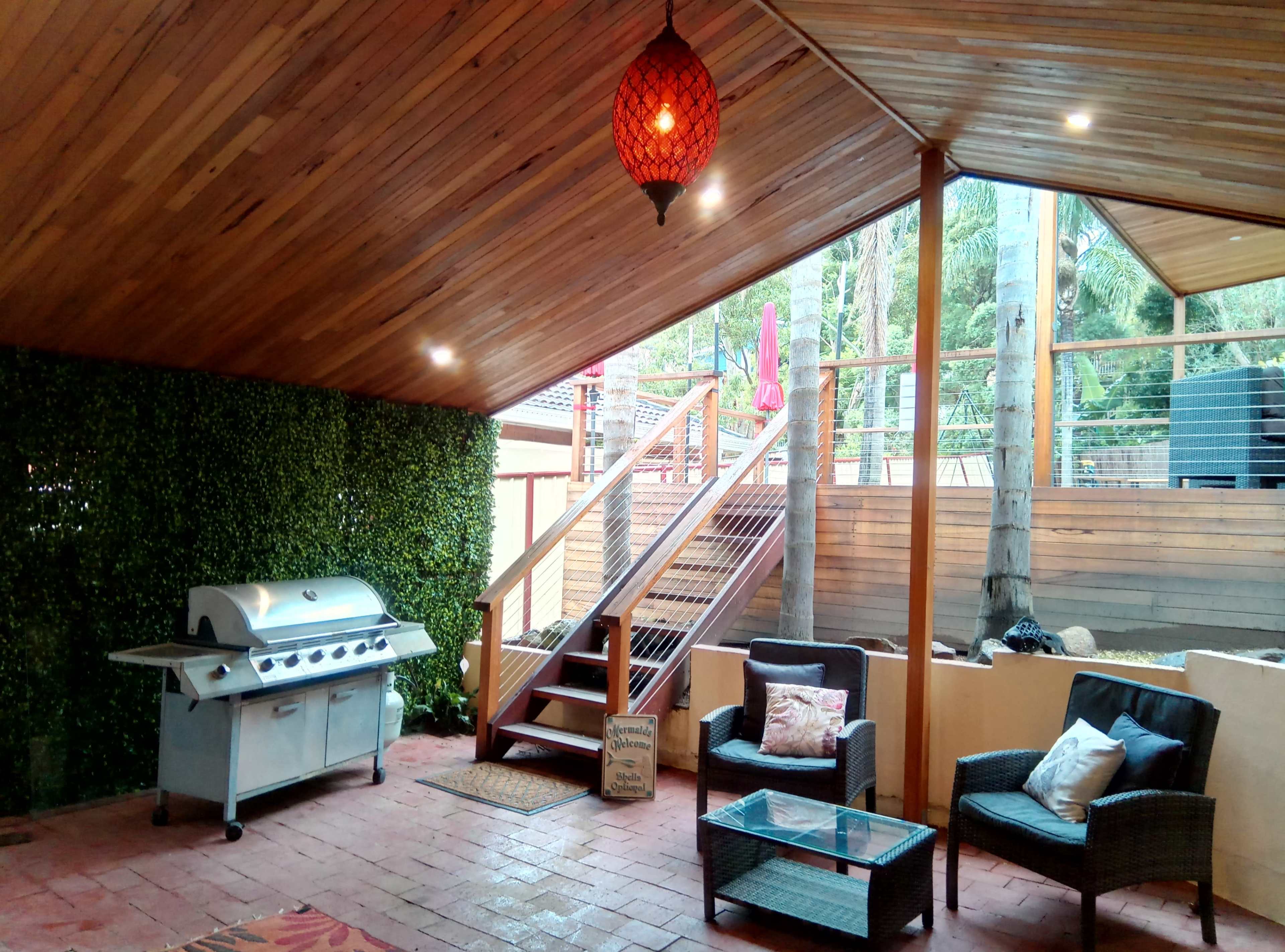 An outdoor patio features a grill, a glass coffee table, two chairs, and wooden stairs leading to a higher deck, all under a wooden ceiling with a hanging light.