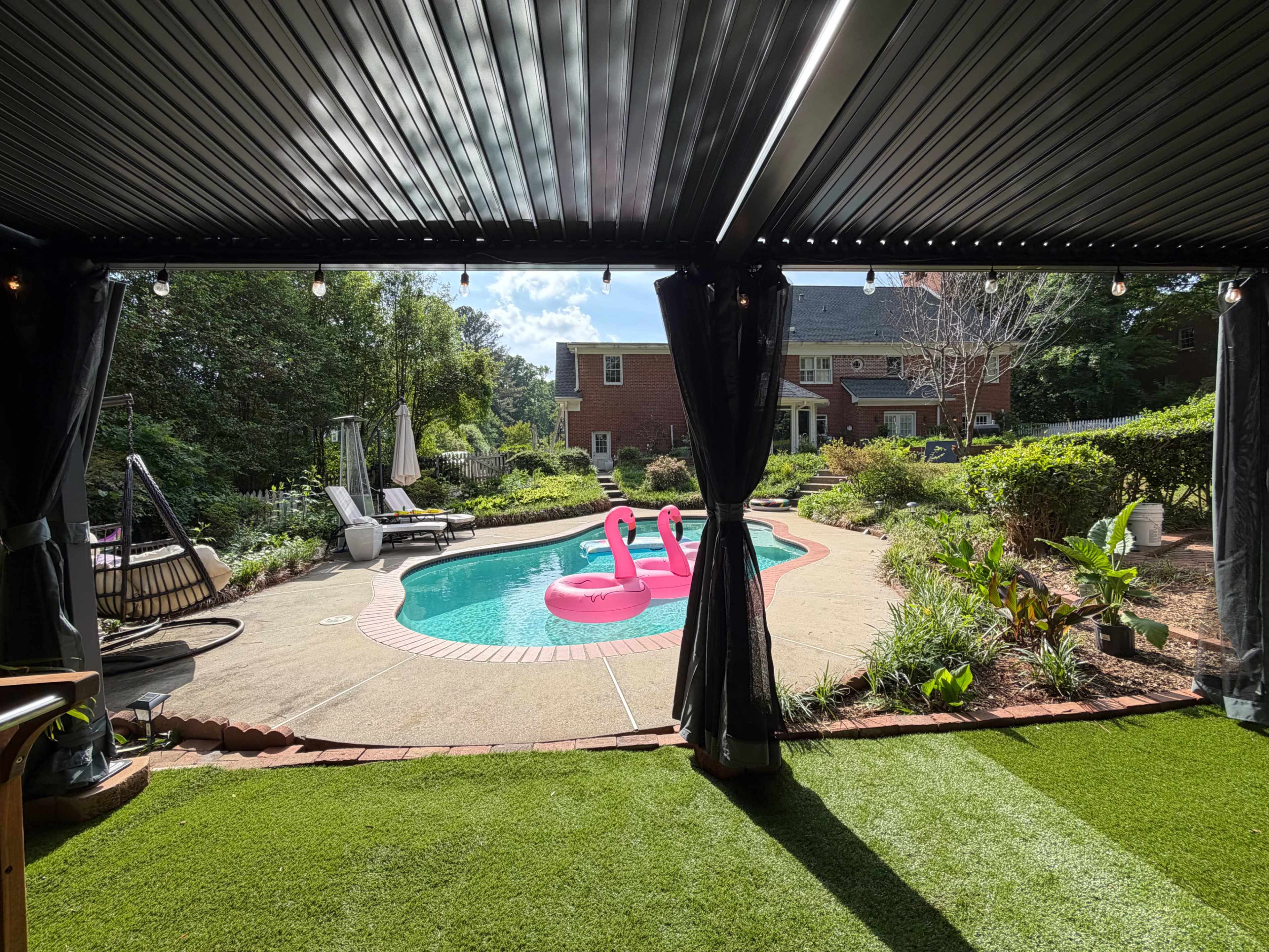 The image shows a backyard with a swimming pool featuring two pink inflatable flamingos, surrounded by green landscaping and outdoor furniture.
