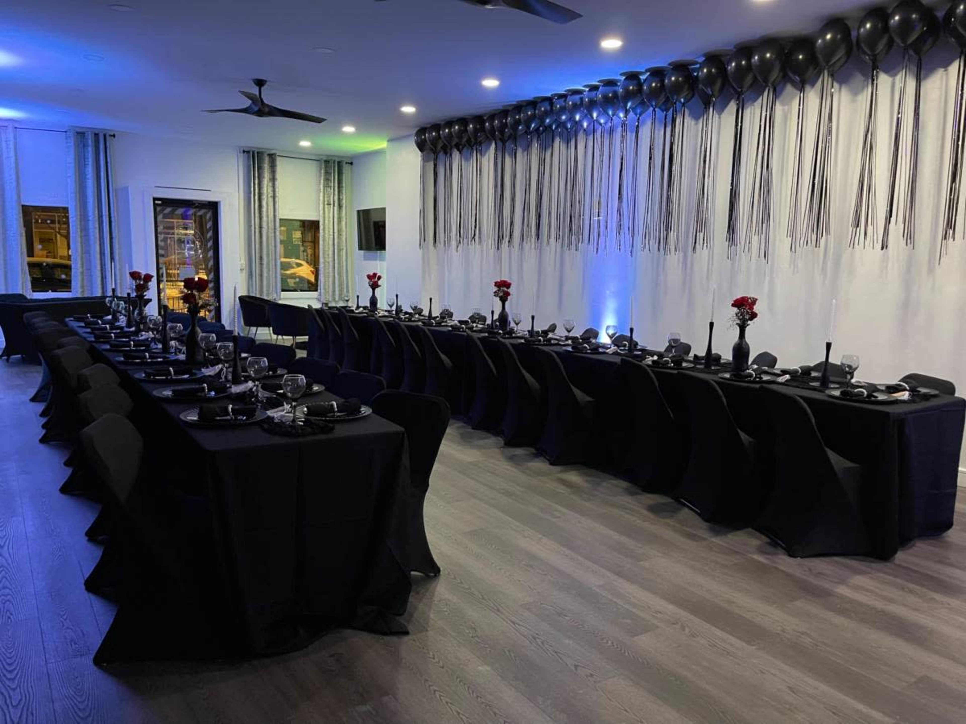 A reception area is set with two long tables covered in black tablecloths, featuring floral centerpieces and adorned with black balloons and streamers.