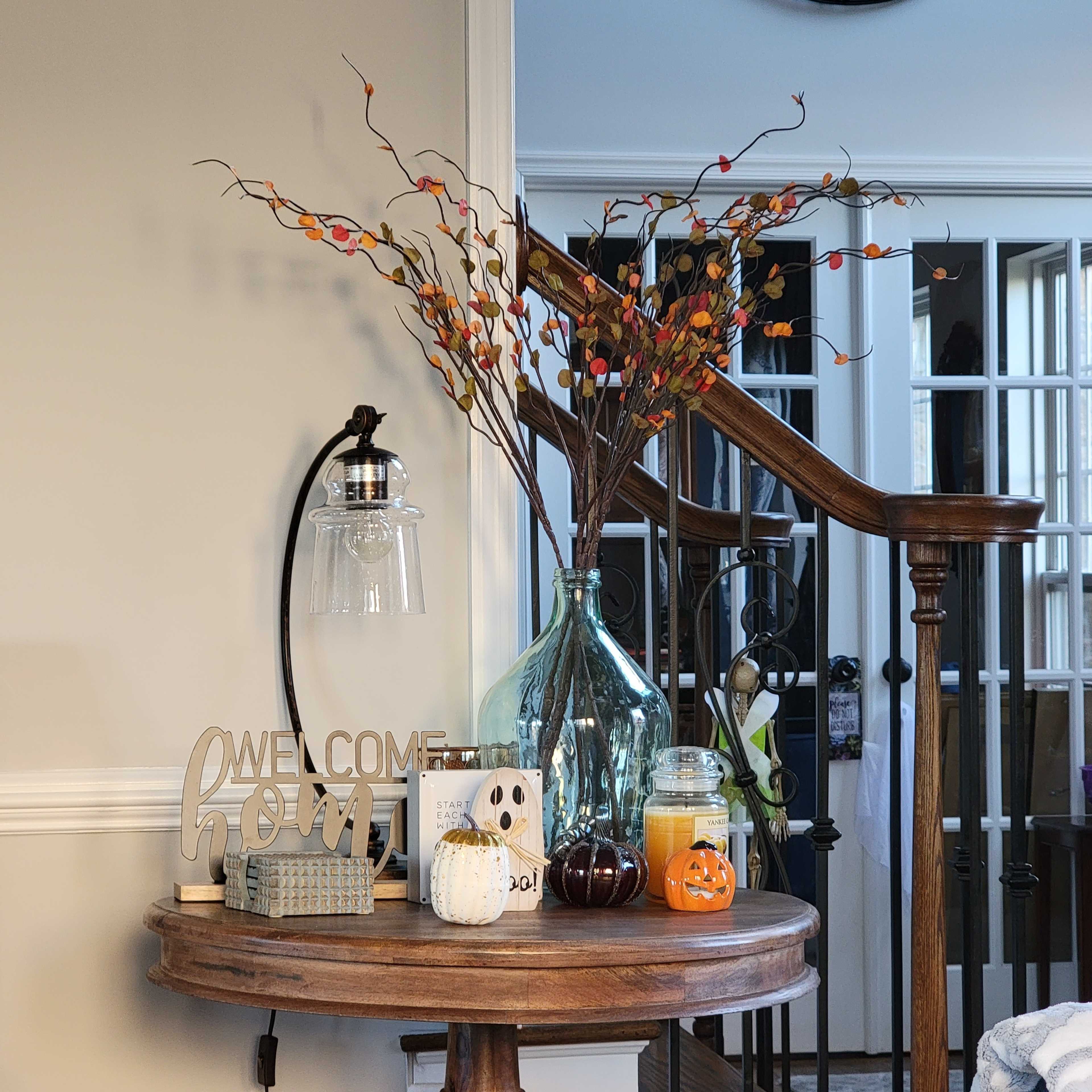 A round table in an entryway features a decorative arrangement with a large glass vase, a lamp, candles, a small pumpkin, and a sign that says "Welcome Home."
