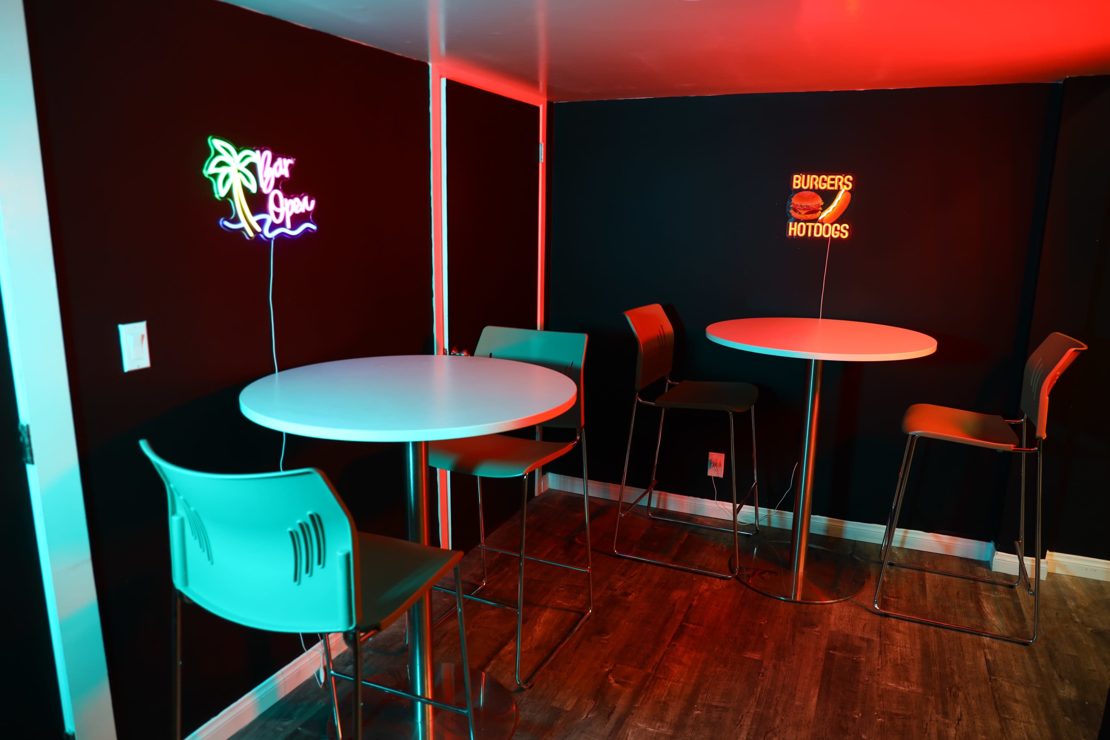 The image shows a small dining area with two round tables and four metal chairs, illuminated by neon signs that read "Bar Love" and "Burgers & Hotdogs."