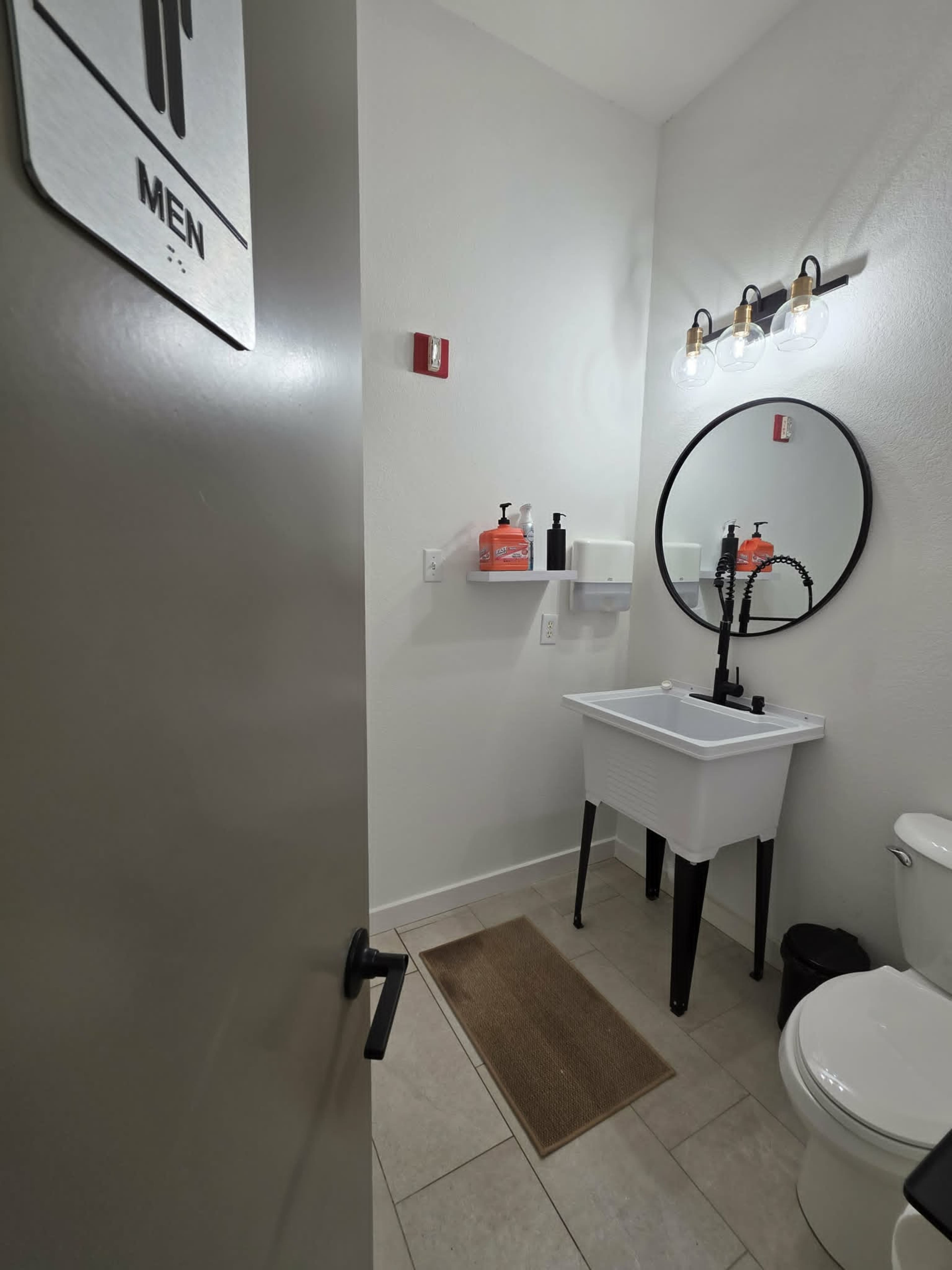 The image shows a men's bathroom with a sink, a round mirror, and a toilet, along with hand sanitizer and paper towel dispensers on the wall.
