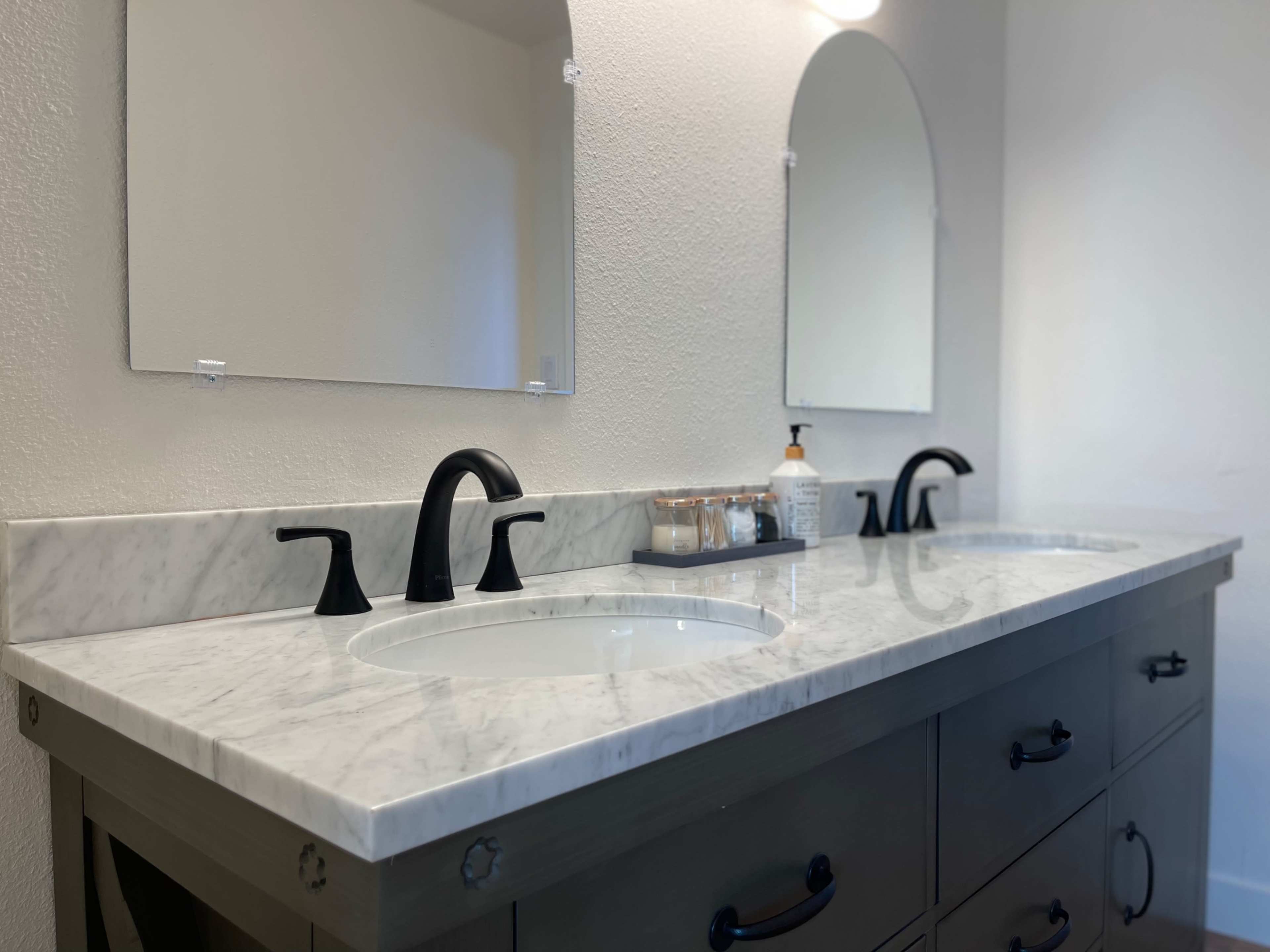 The image shows a bathroom vanity with a double sink, a white marble countertop, and black faucets, accompanied by two mirrors and a bottle on the counter.