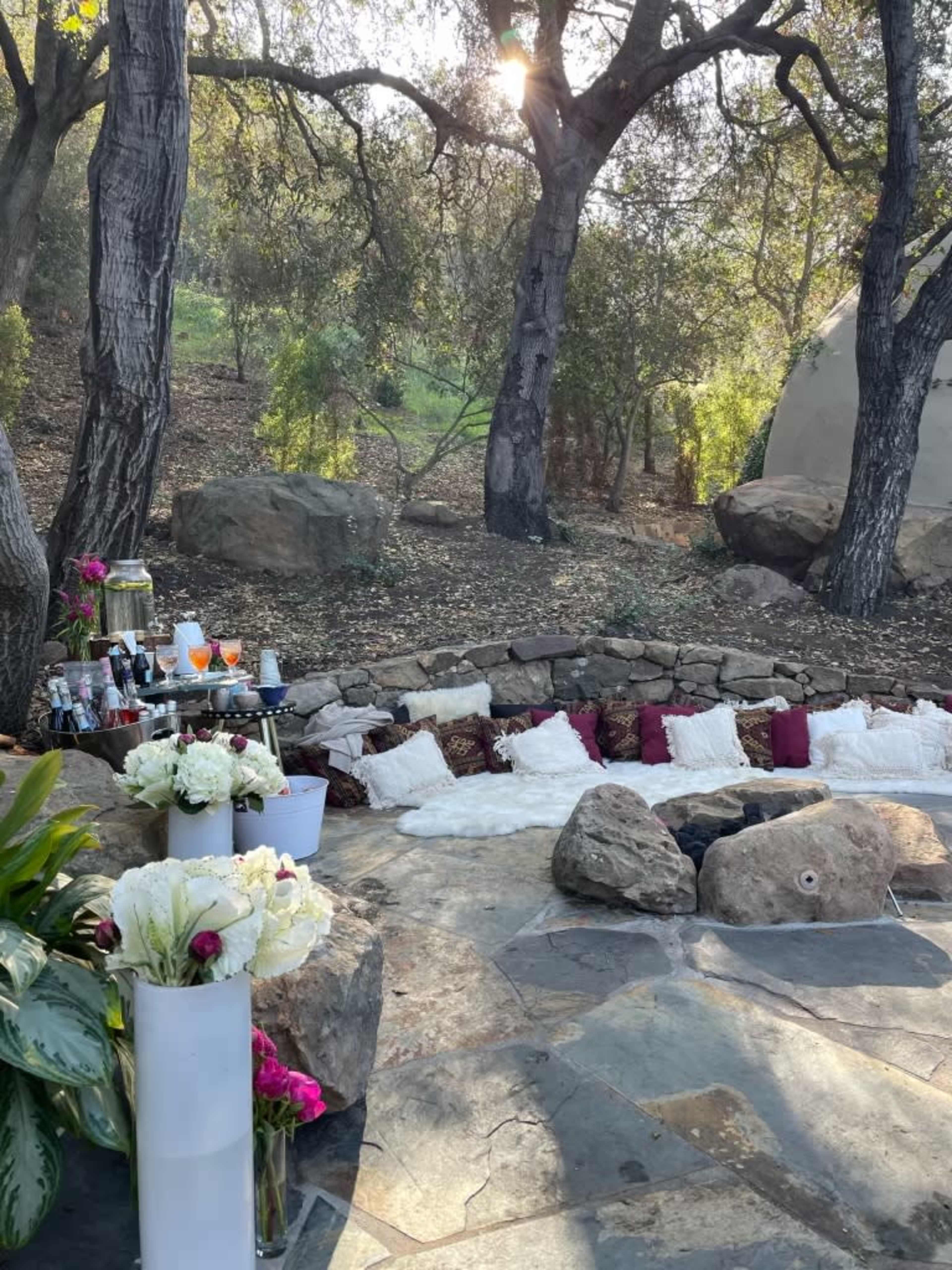 The scene features a rustic outdoor setting with a row of cushions arranged on stone tiles, surrounded by trees and large rocks, alongside a small table with drinks and flowers.