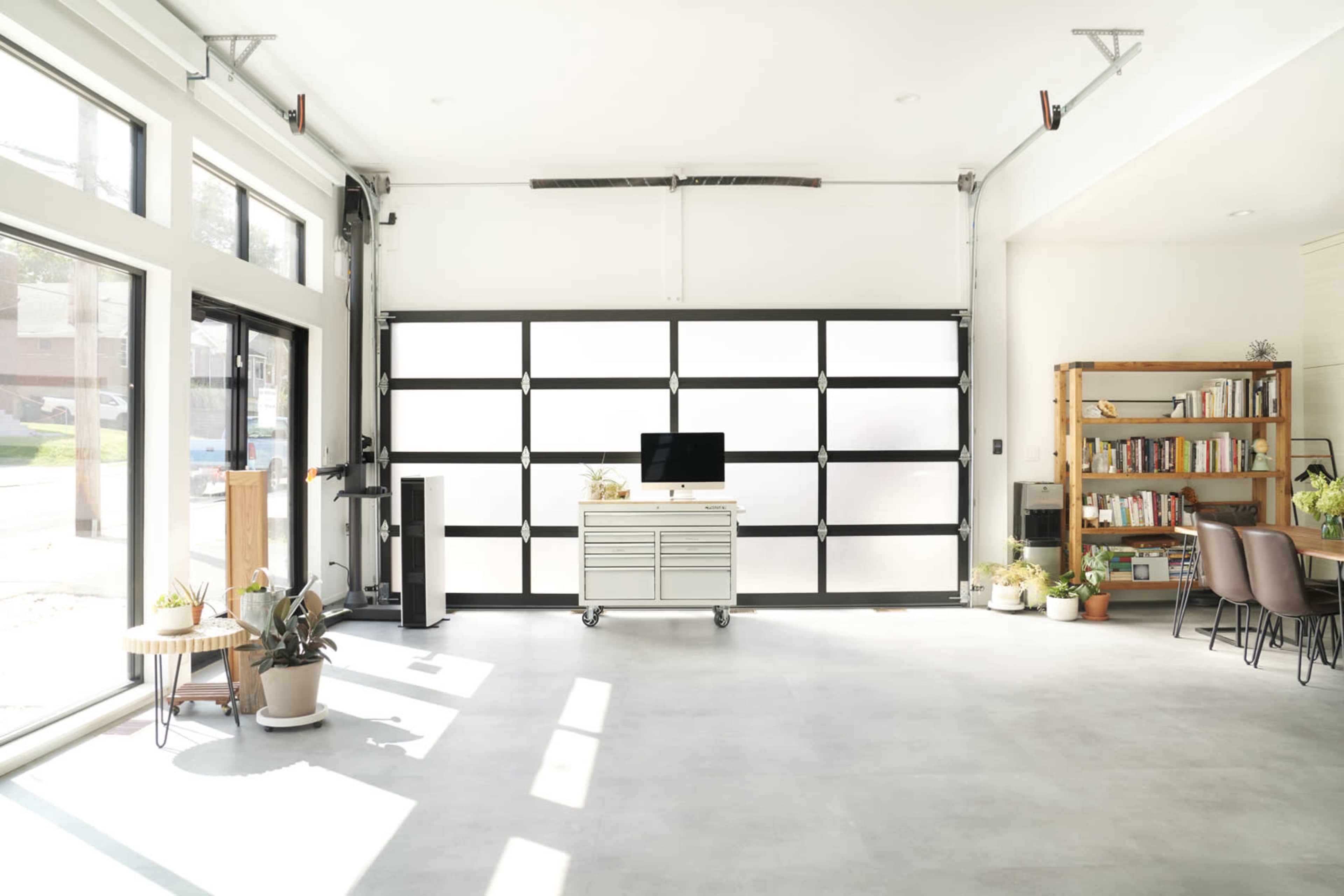 The image shows a modern garage with a large glass door, a workbench on wheels, a television, a bookshelf filled with books, and potted plants.