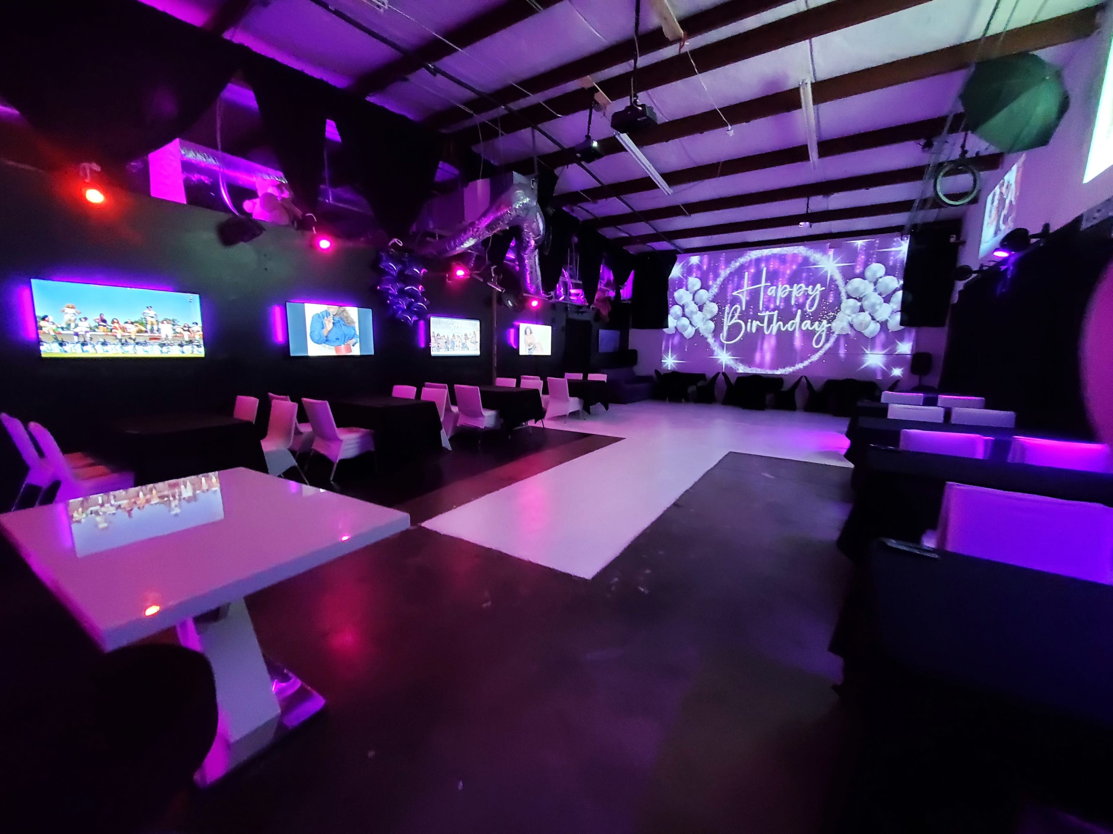 The image shows a spacious event venue with purple and pink lighting, decorated with balloons and featuring screens displaying birthday messages.