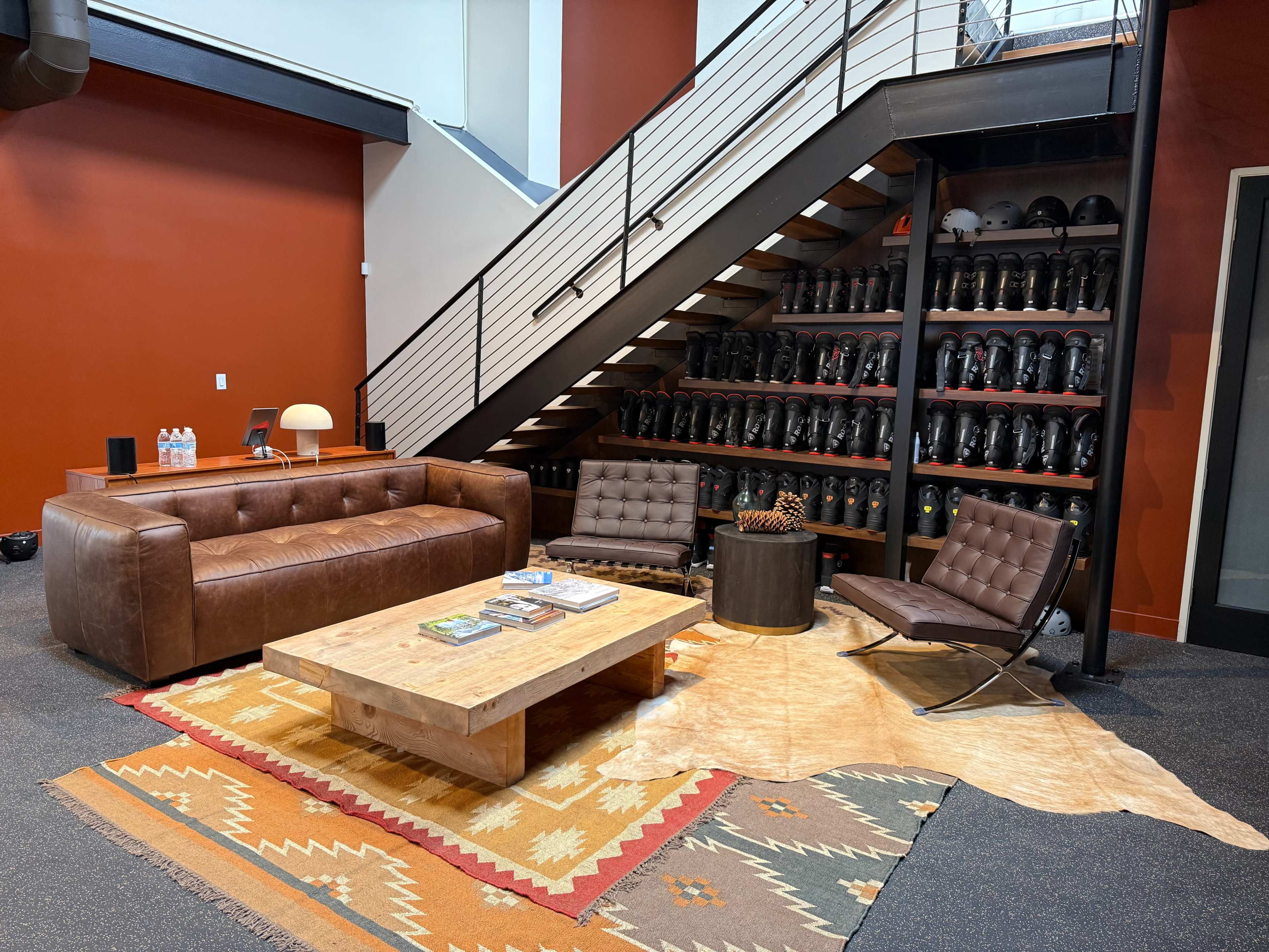 The image shows a modern lounge area with a brown leather sofa, a wooden coffee table, and a vintage rug, situated next to a staircase and a wall lined with black helmets.