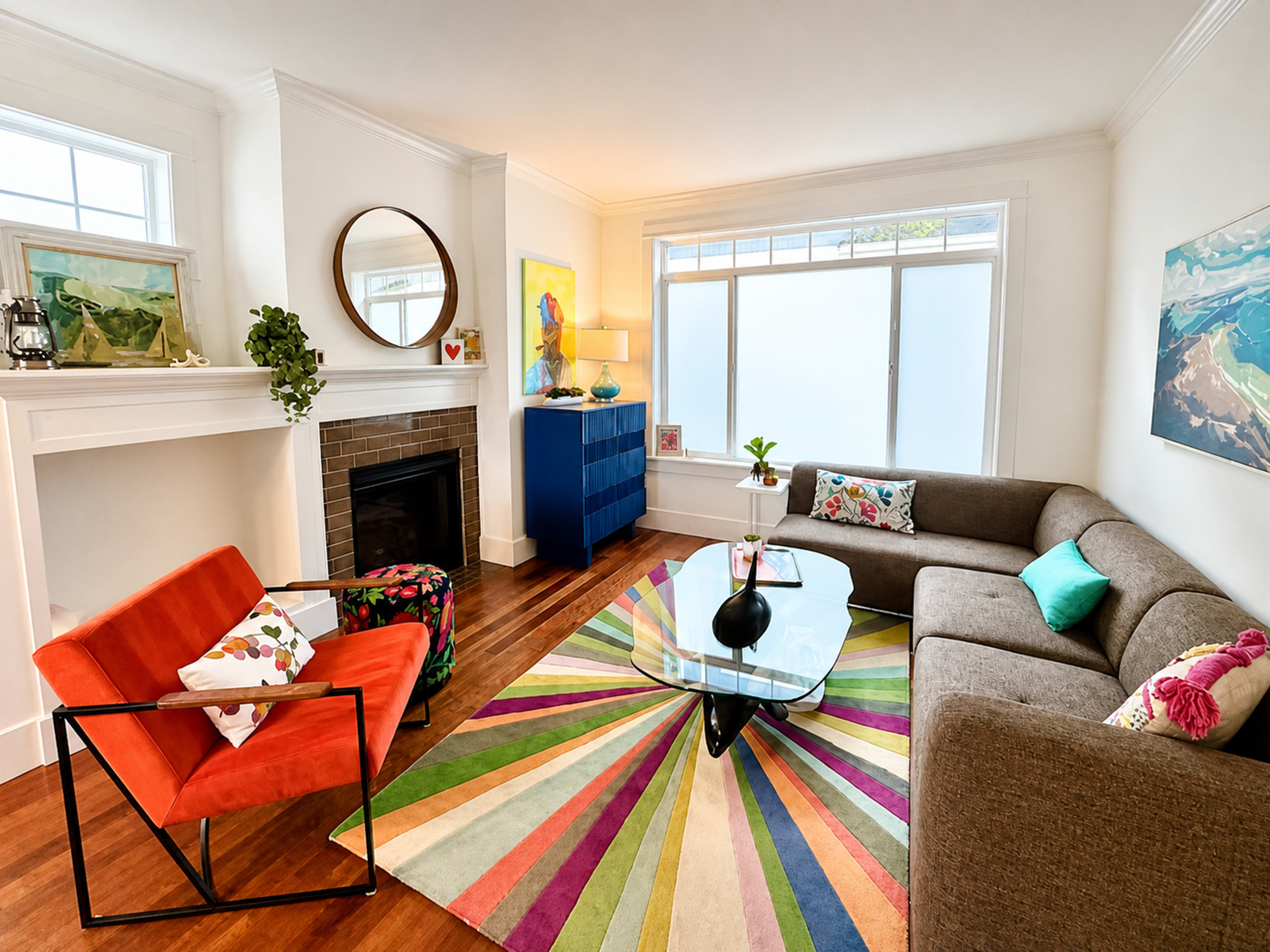 The image shows a modern living room featuring a colorful striped rug, a glass coffee table, a gray sofa, an orange chair, and a blue cabinet near a window.