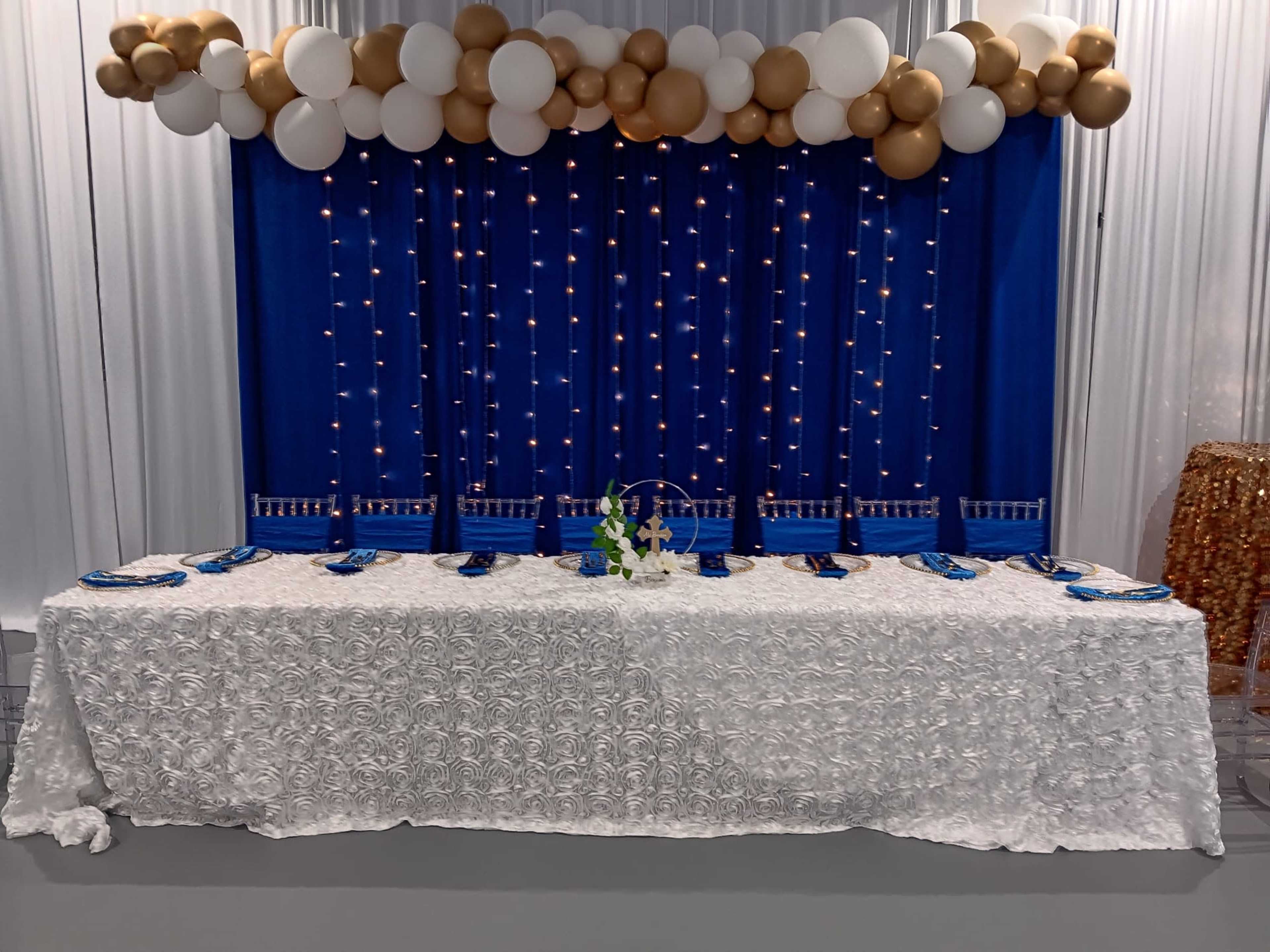 The image shows a decorated table with a white textured tablecloth, blue napkins, and a floral centerpiece, set against a backdrop of blue fabric and golden and white balloons.