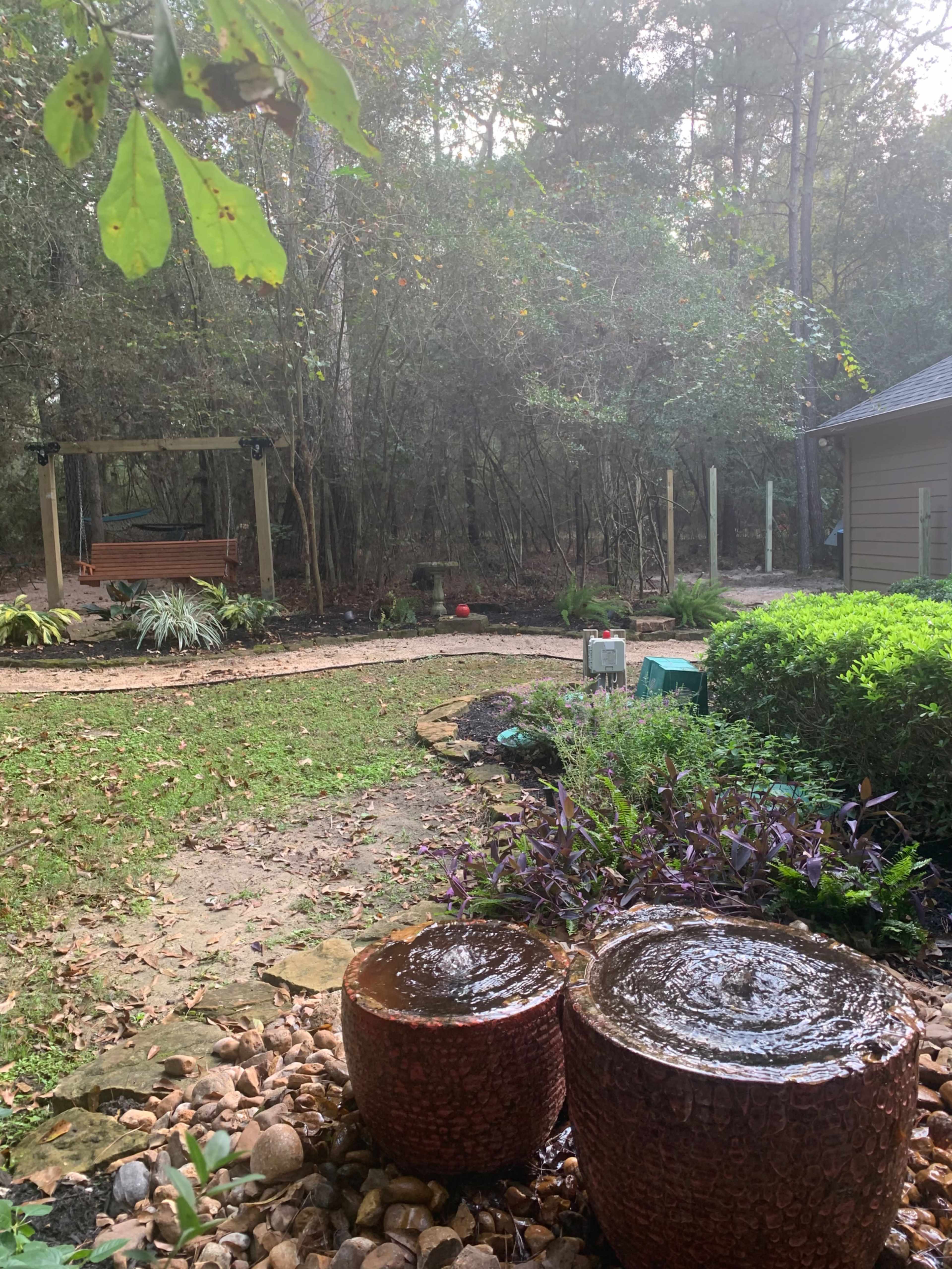 The scene shows a backyard with two stone water features, a swing in the background, and a path lined by greenery.