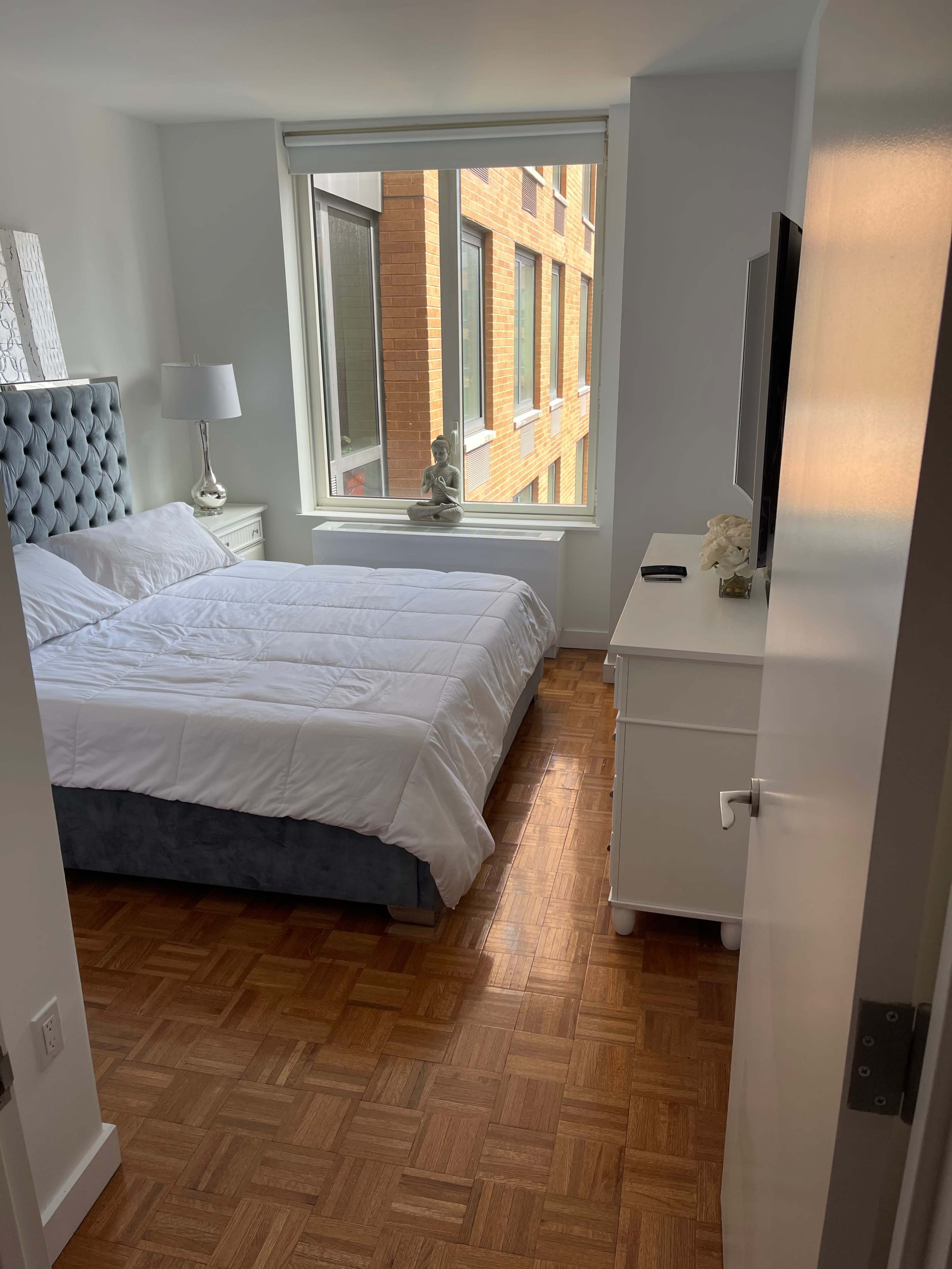 A neatly arranged bedroom features a bed with a gray headboard, a white comforter, a nightstand with a lamp, and a window showing a brick building outside.