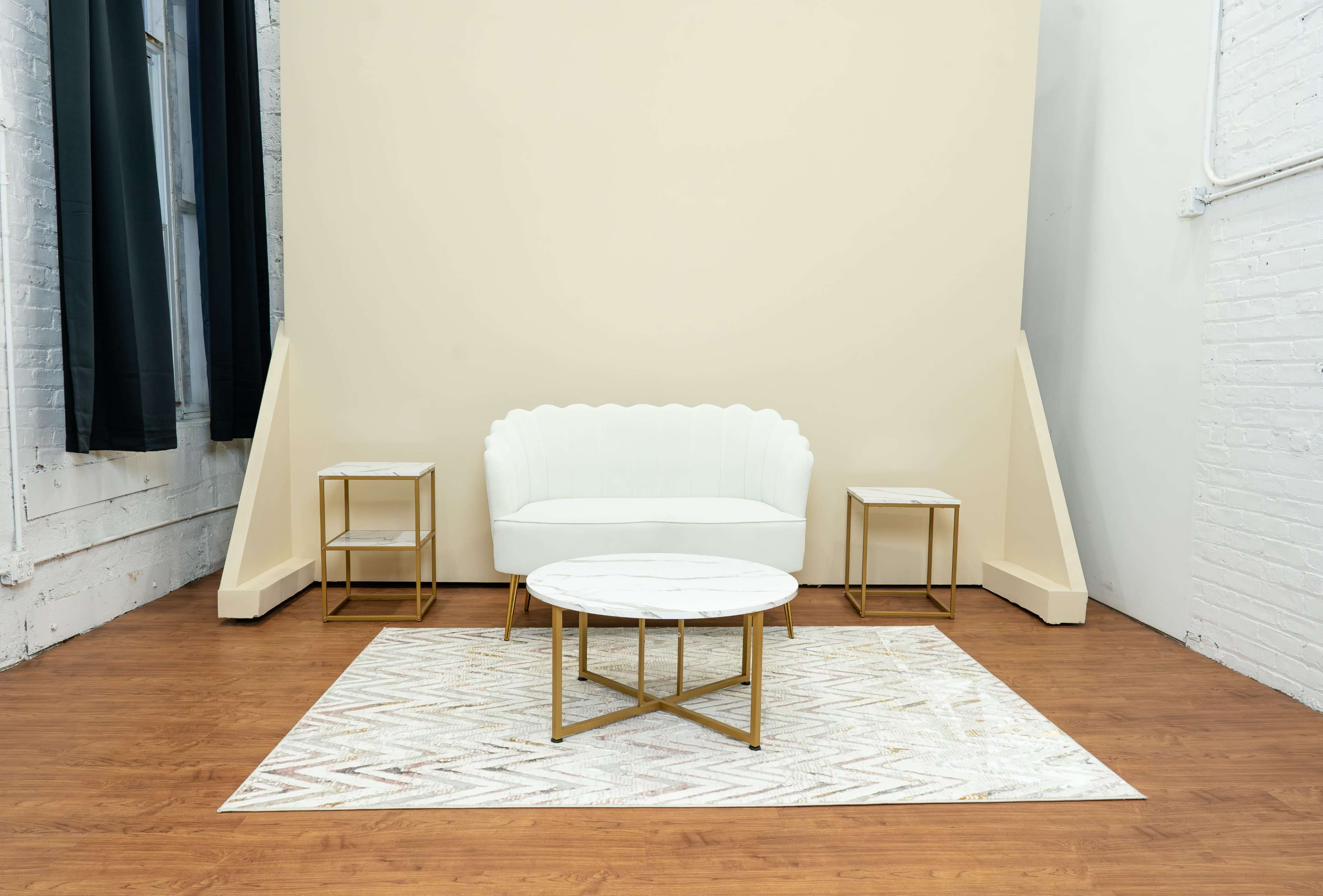 A white scalloped sofa and two side tables are arranged on a patterned rug in a minimalist interior with a light-colored backdrop.