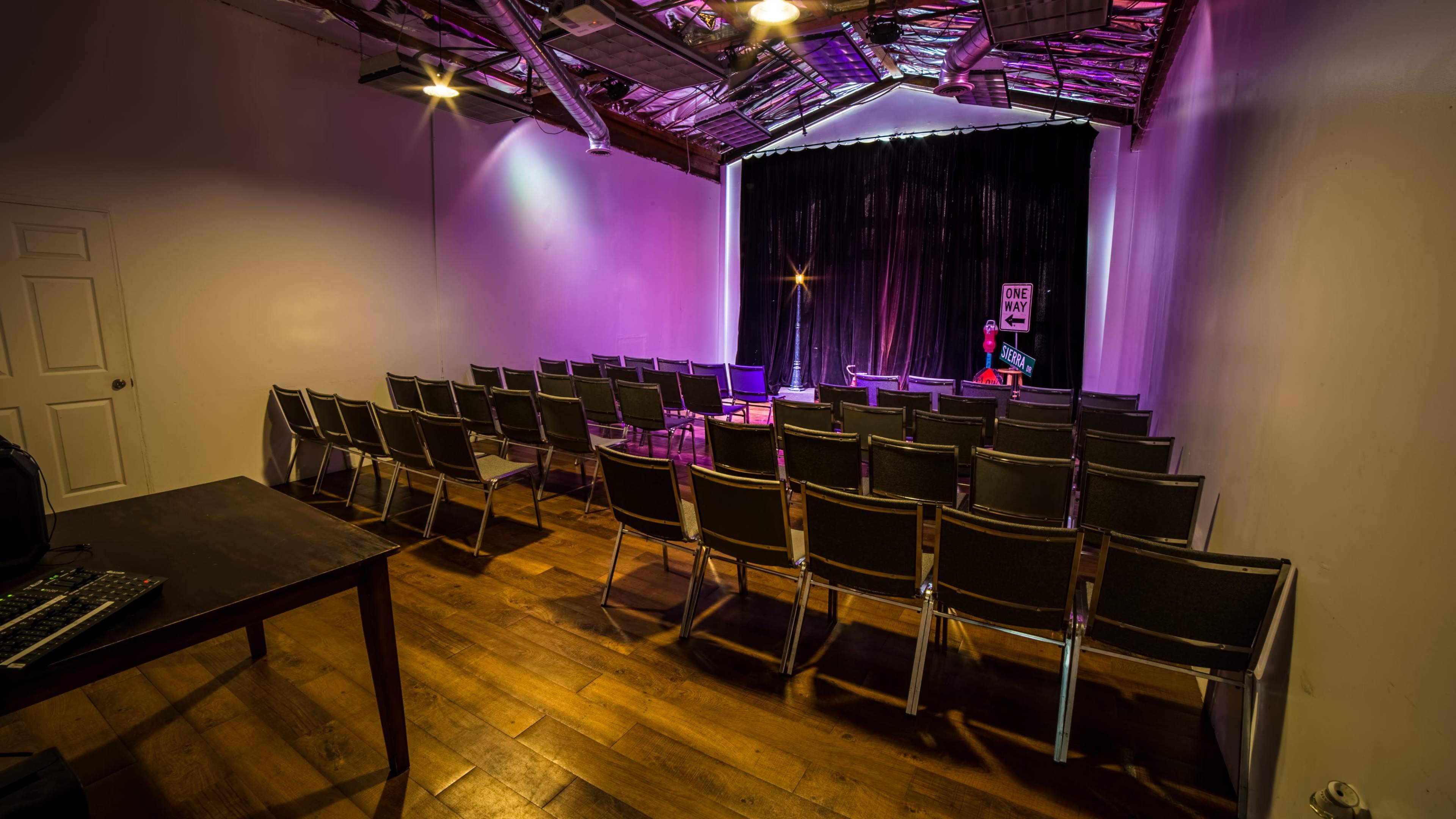 A dimly lit performance space features rows of empty chairs facing a stage with a black curtain and a spotlight.