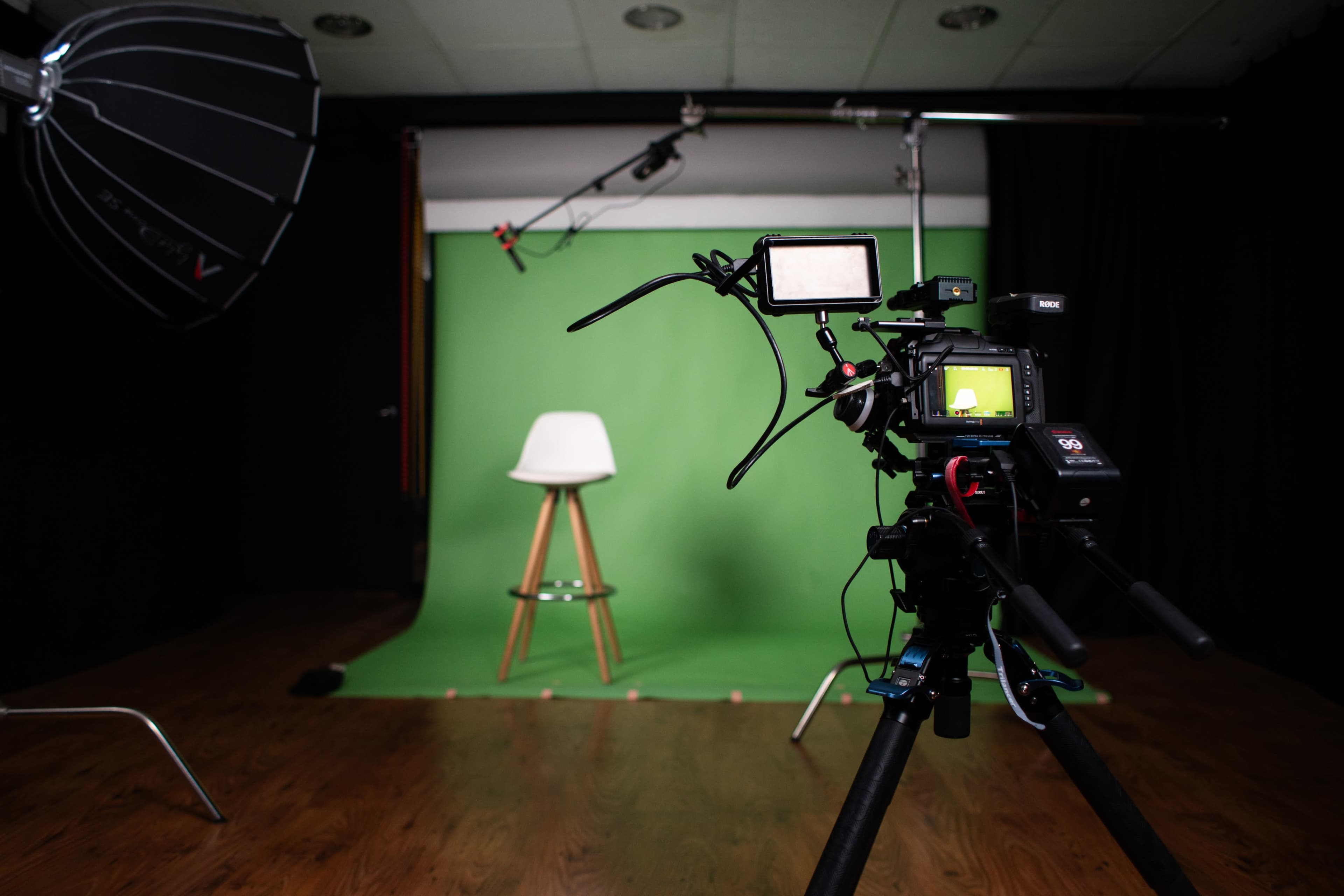 A camera is positioned in front of a green screen with a white stool placed on a wooden floor in a studio setting.