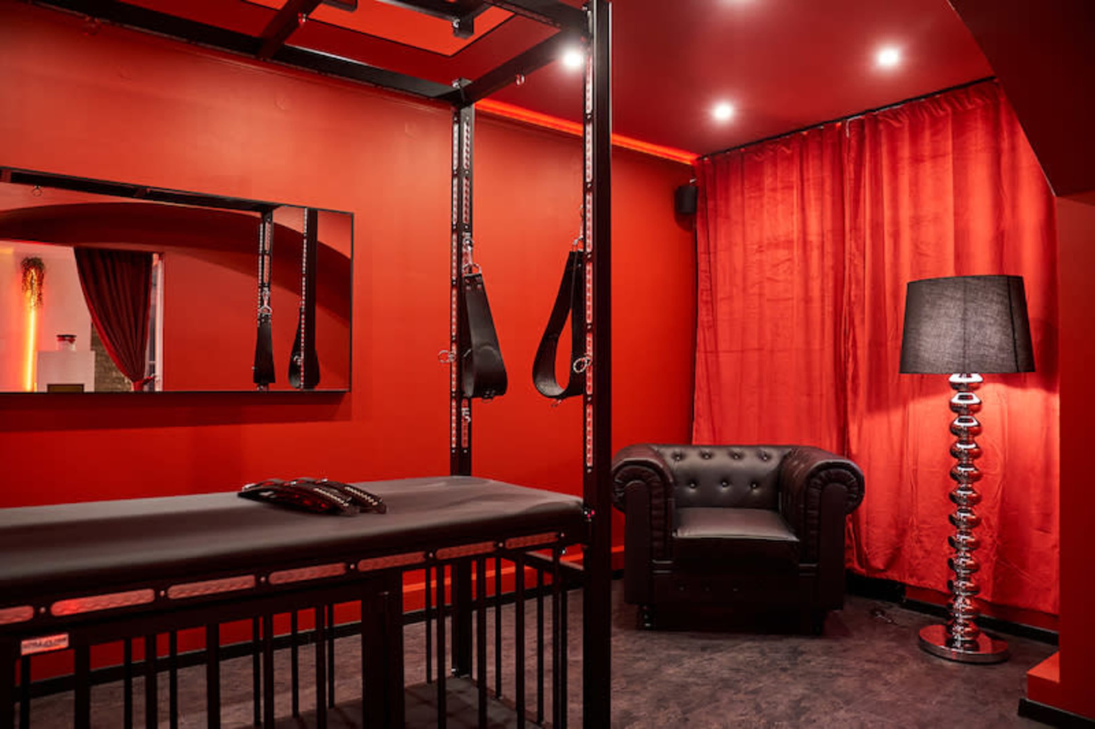 The image shows a room with red walls featuring a metal restraint frame, a black padded bench, a mirror, a leather couch, and a lamp.