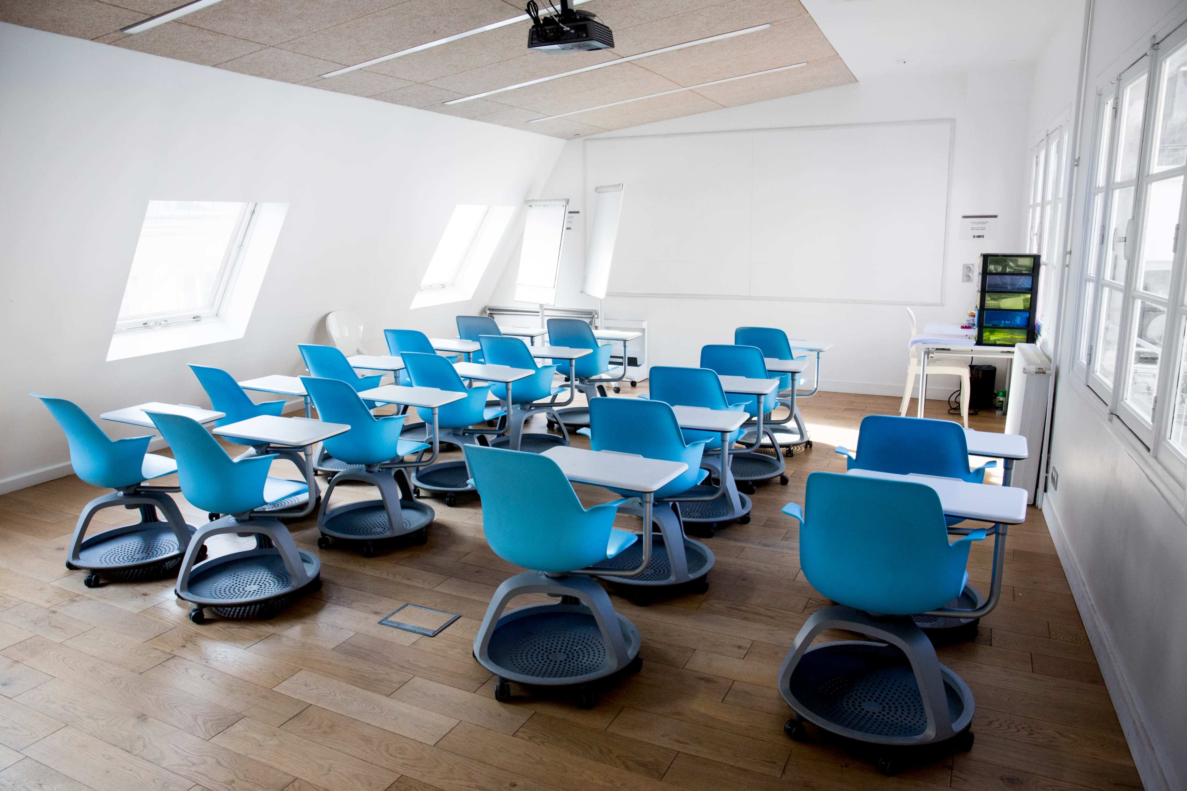A classroom is set up with blue rolling chairs arranged around small desks, and large windows allowing natural light to fill the space.