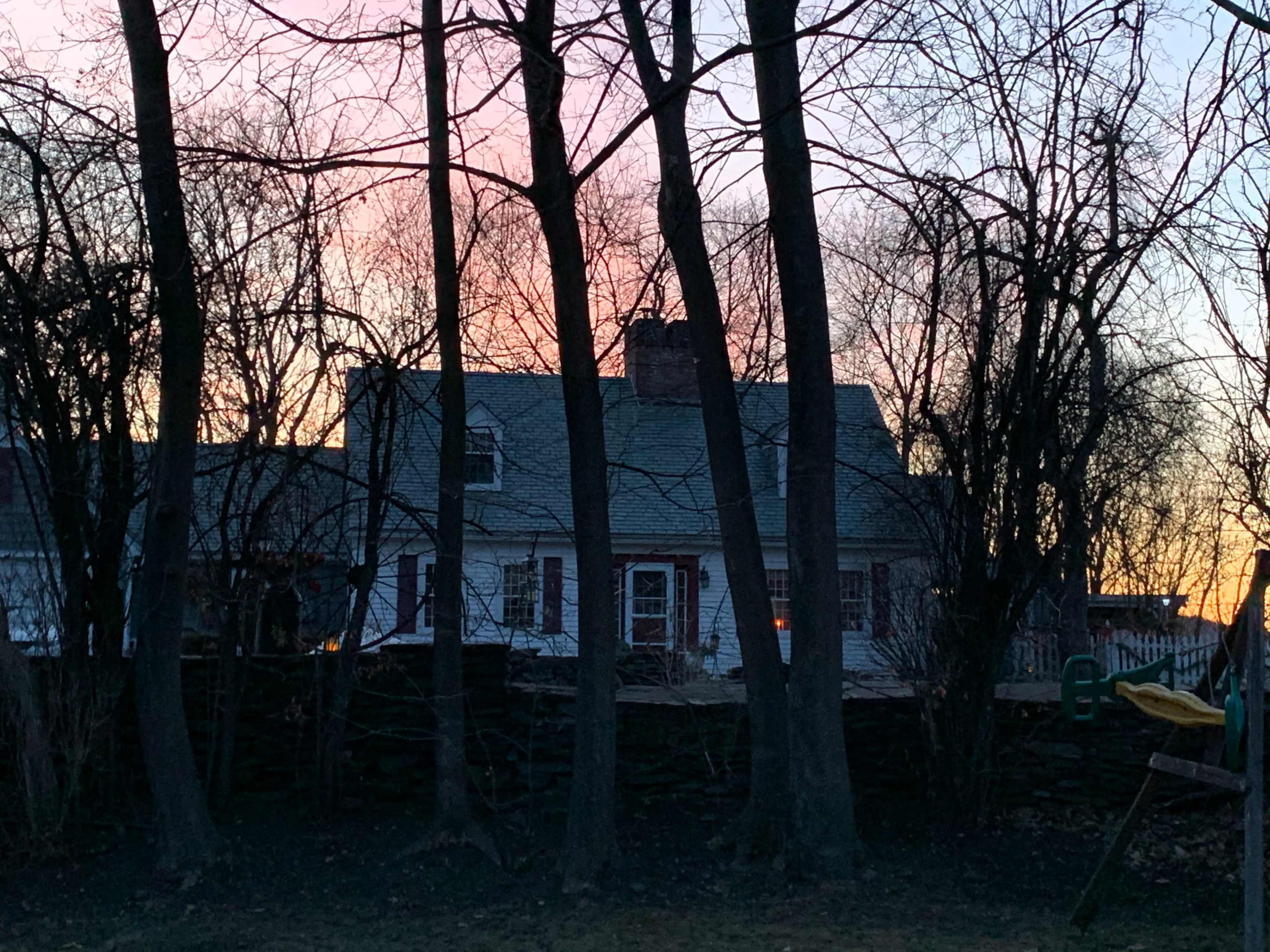A house is partially obscured by trees during a sunset, with a warm glow visible through the windows.
