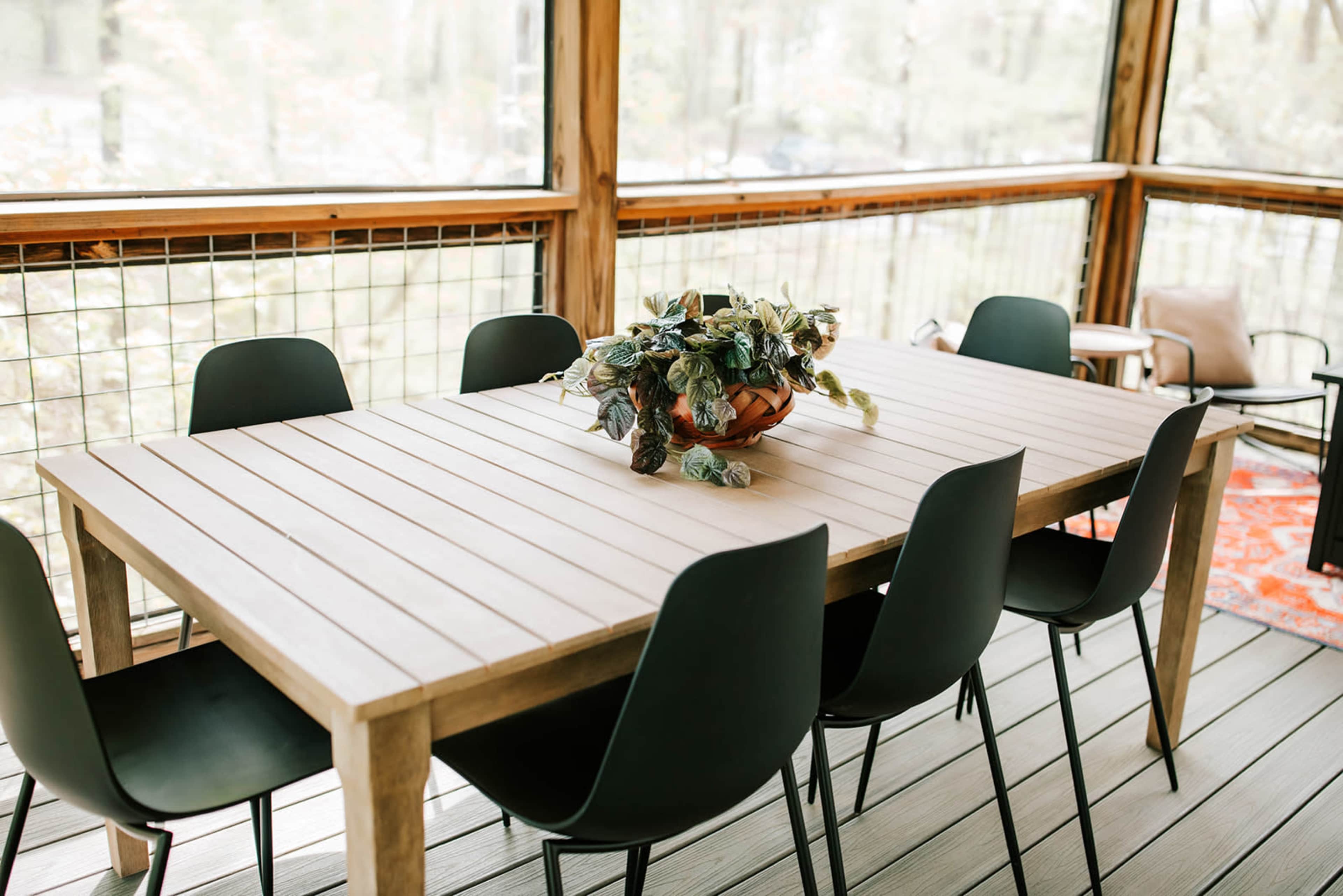 A wooden dining table with a potted plant centerpiece is surrounded by six black chairs in a screened-in porch.