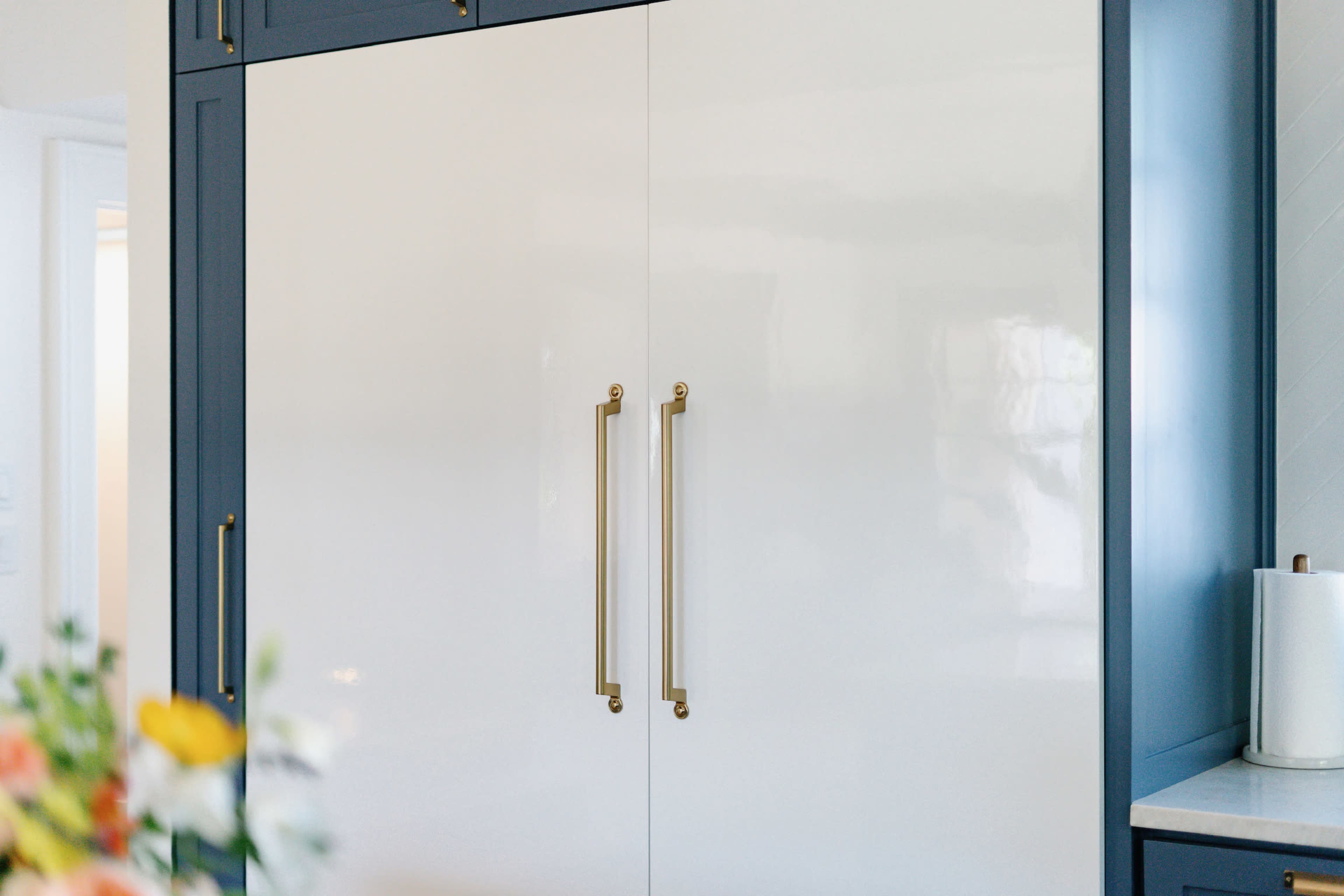 A sleek, modern refrigerator with a glossy white finish and gold handles, set against a blue cabinetry backdrop.