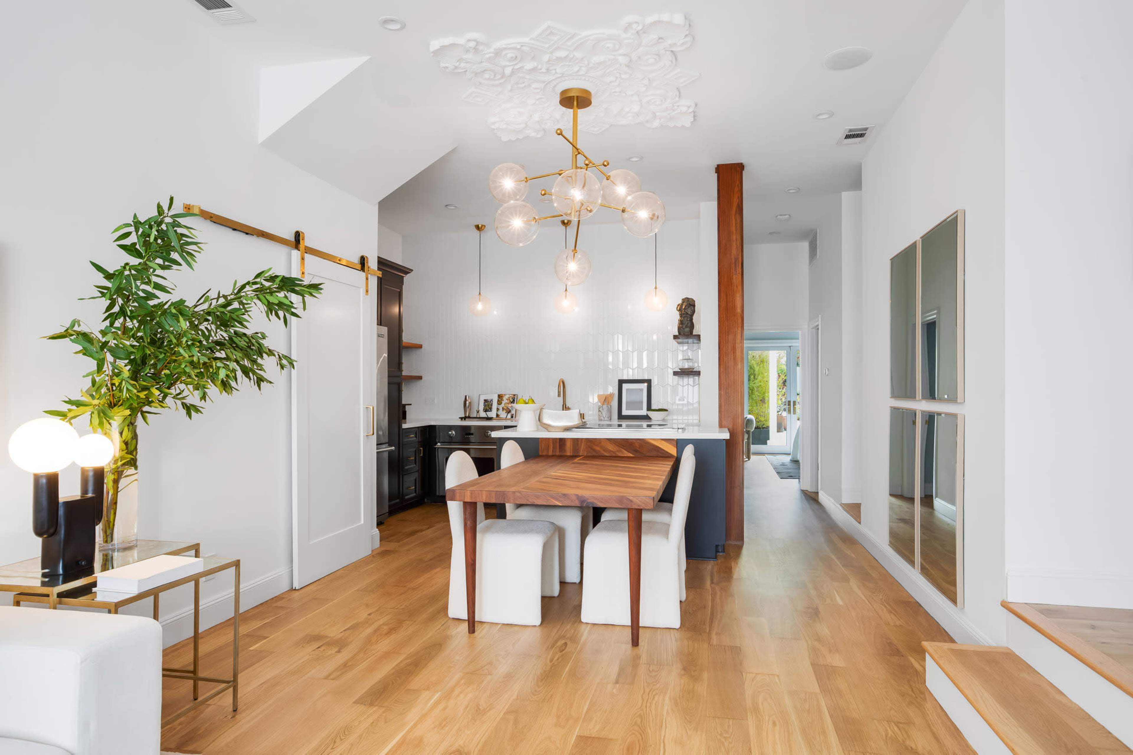 A modern living space features a wooden dining table, white chairs, a decorative chandelier, and a combination of light and dark cabinetry.