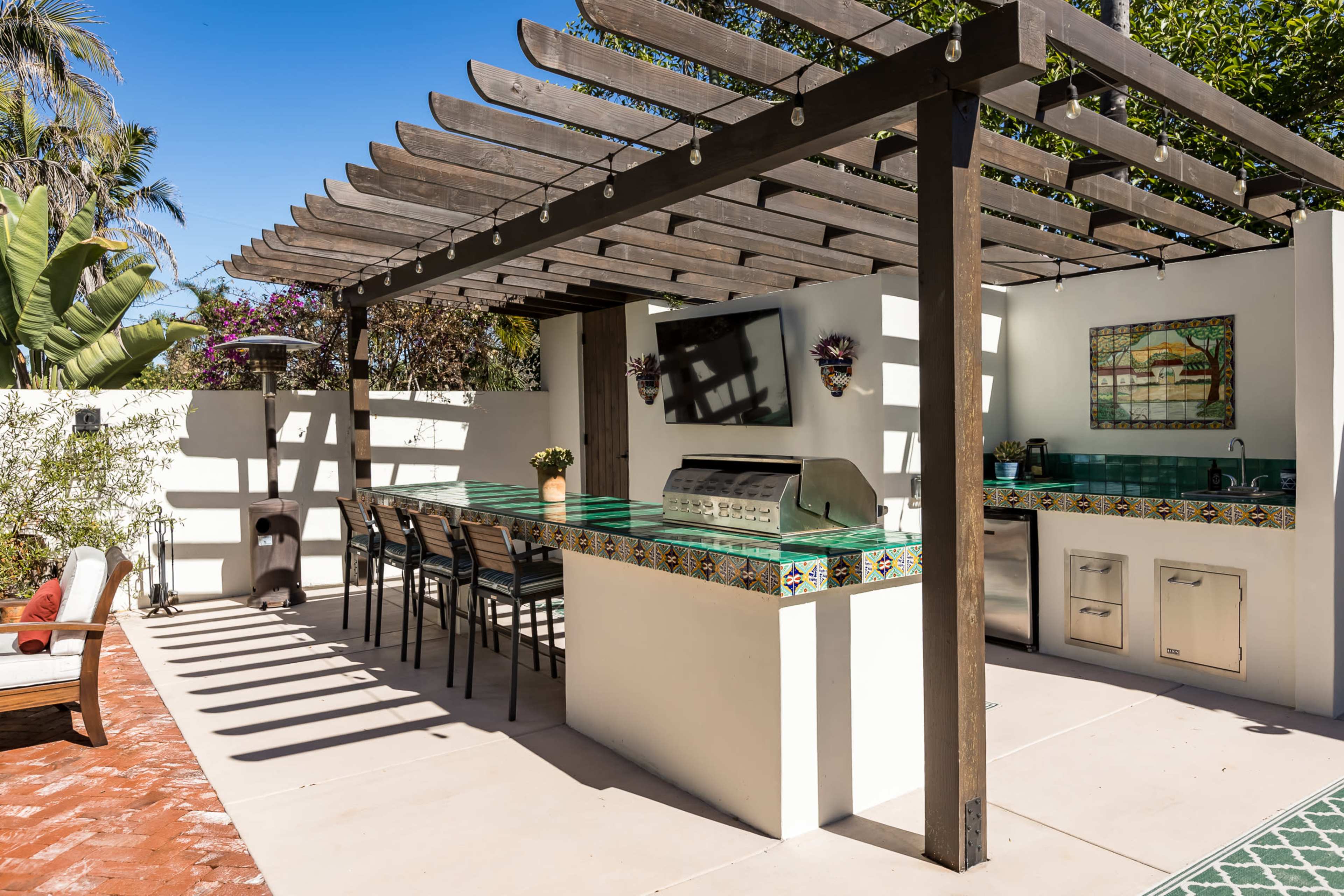 The image shows an outdoor kitchen area with a grill, bar stools, and a decorative tiled counter under a wooden pergola.