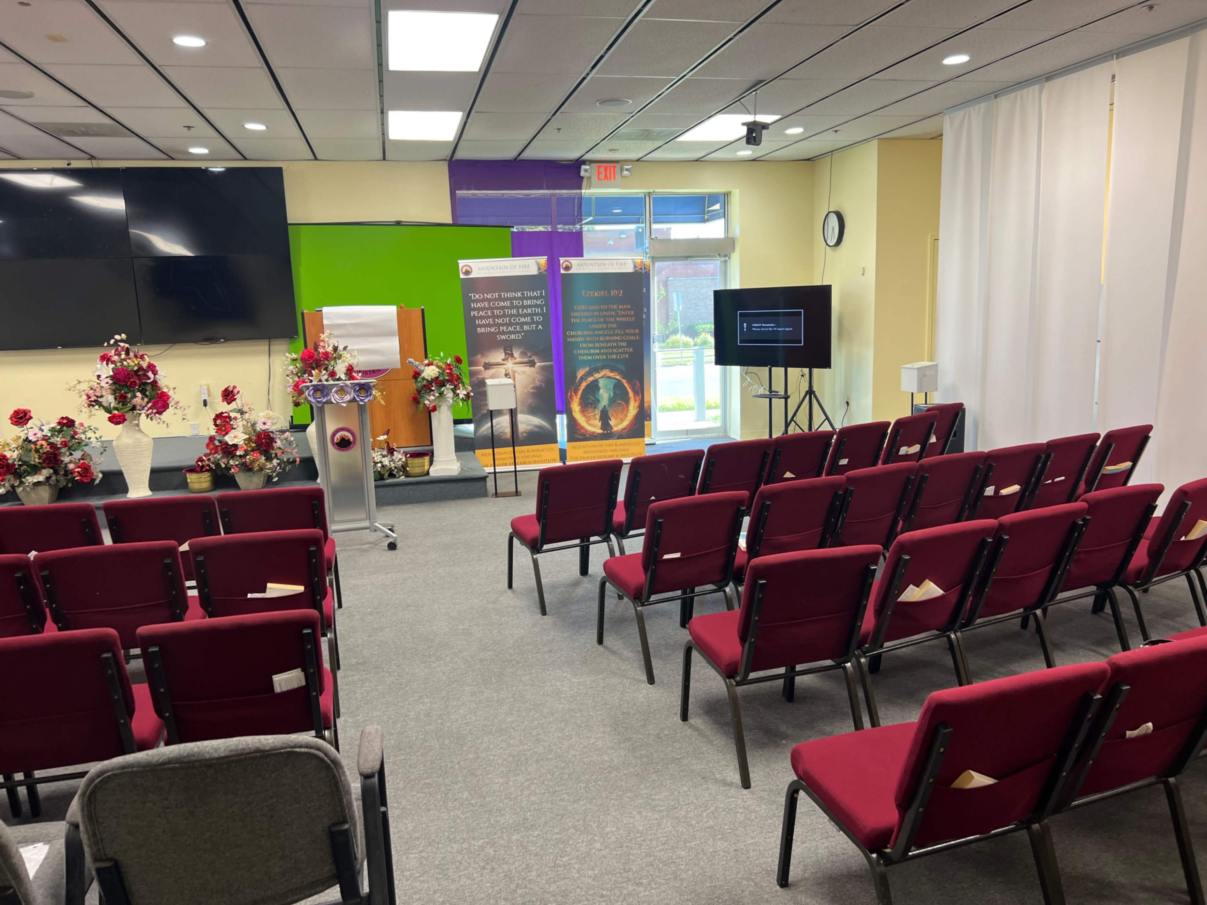 The image shows a seminar room set up with rows of red chairs facing a stage with a podium and floral arrangements on either side, along with screens and banners in the background.