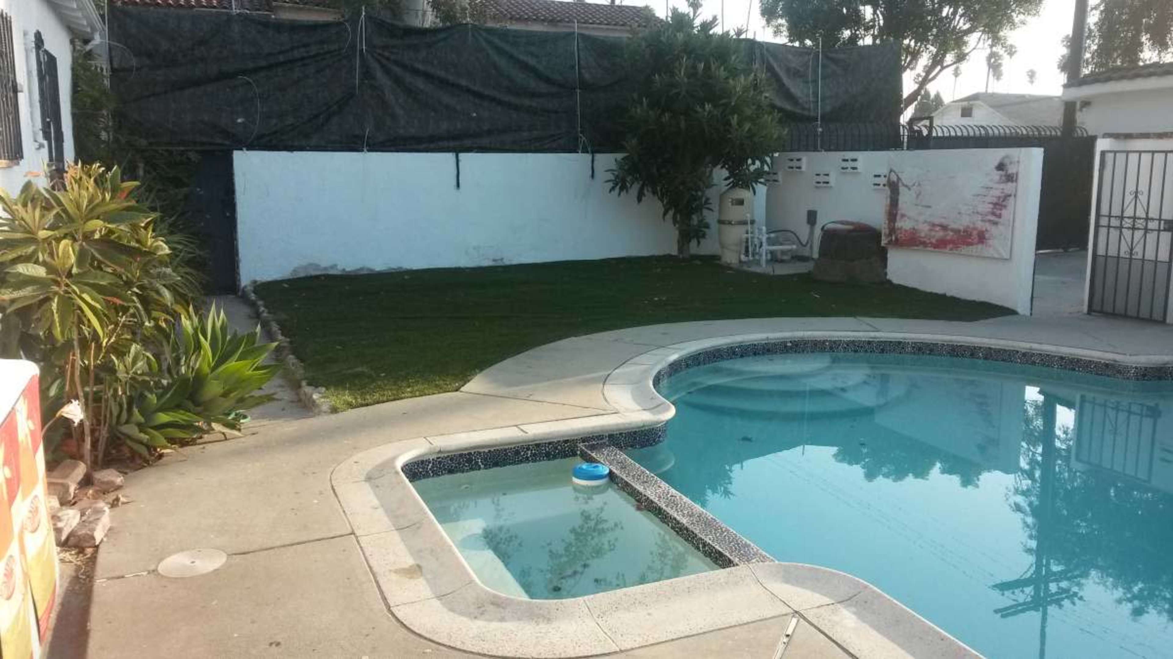 The image shows a backyard with a swimming pool, a small section of grass, and a white wall partially covered by a green tarp.