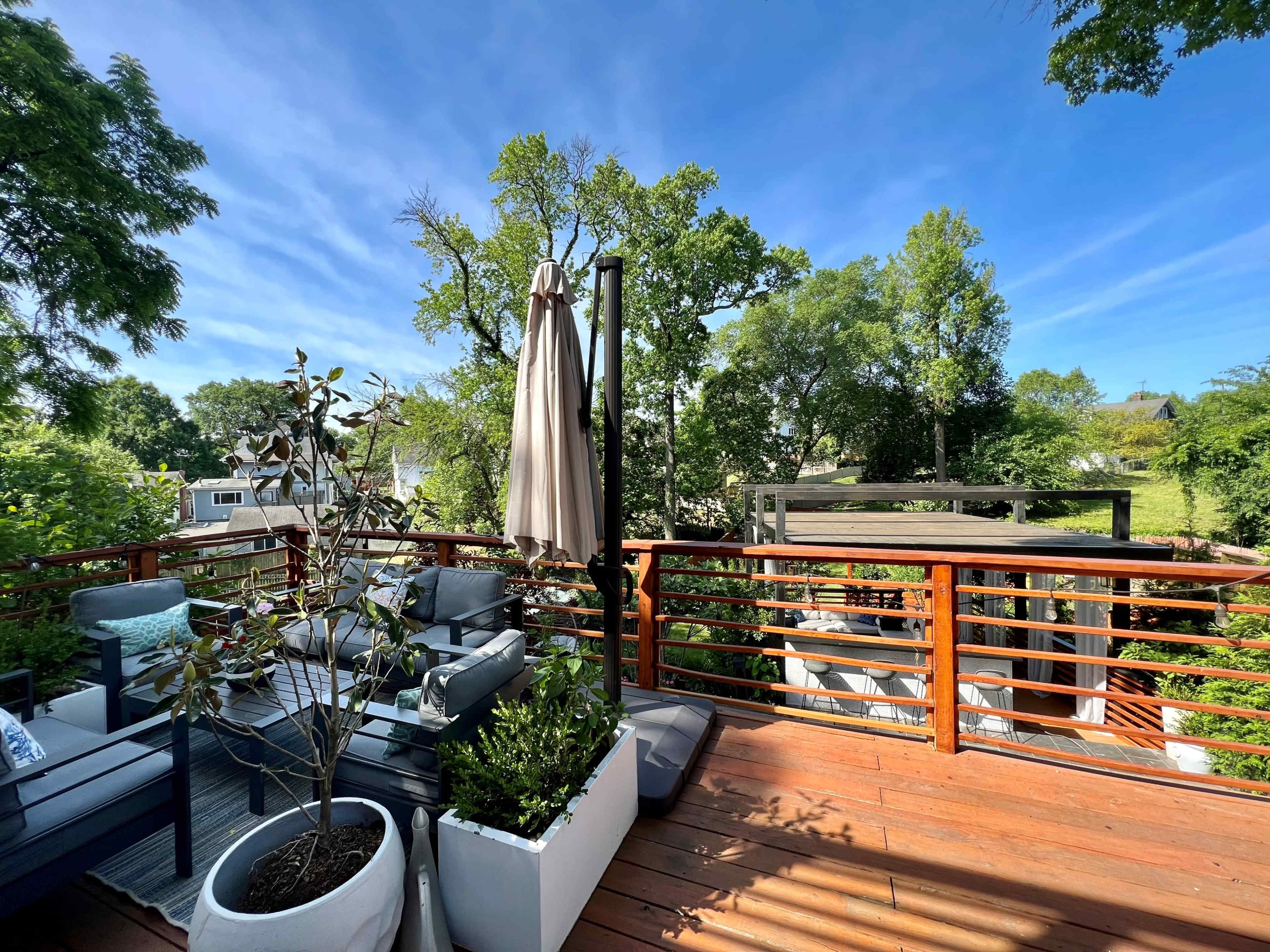 A wooden deck features modern seating arrangements, planters, and a large umbrella among greenery and trees.