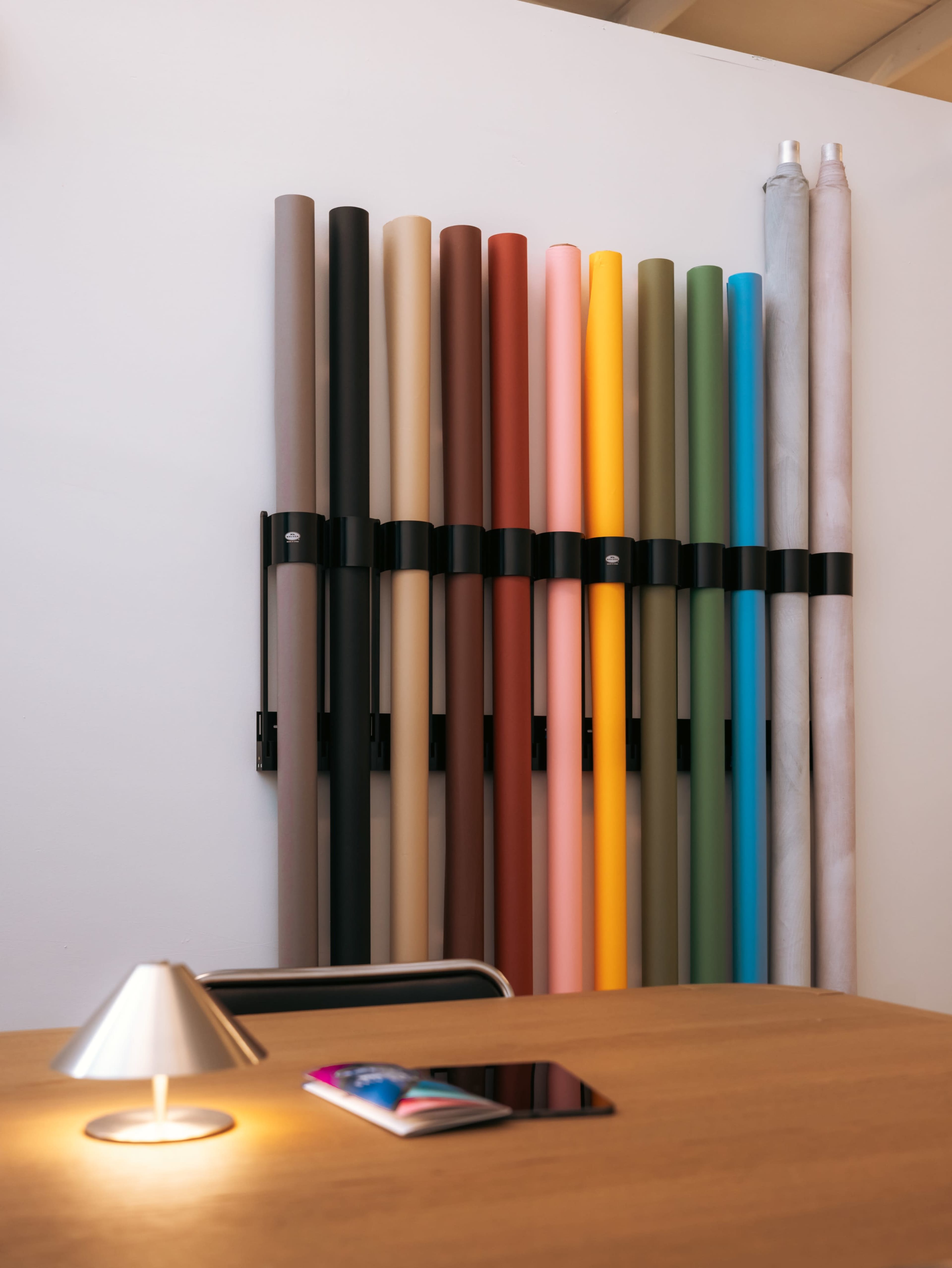 The image shows a collection of neatly arranged rolls of paper in various colors mounted on a wall beside a wooden table with a small lamp and a tablet.
