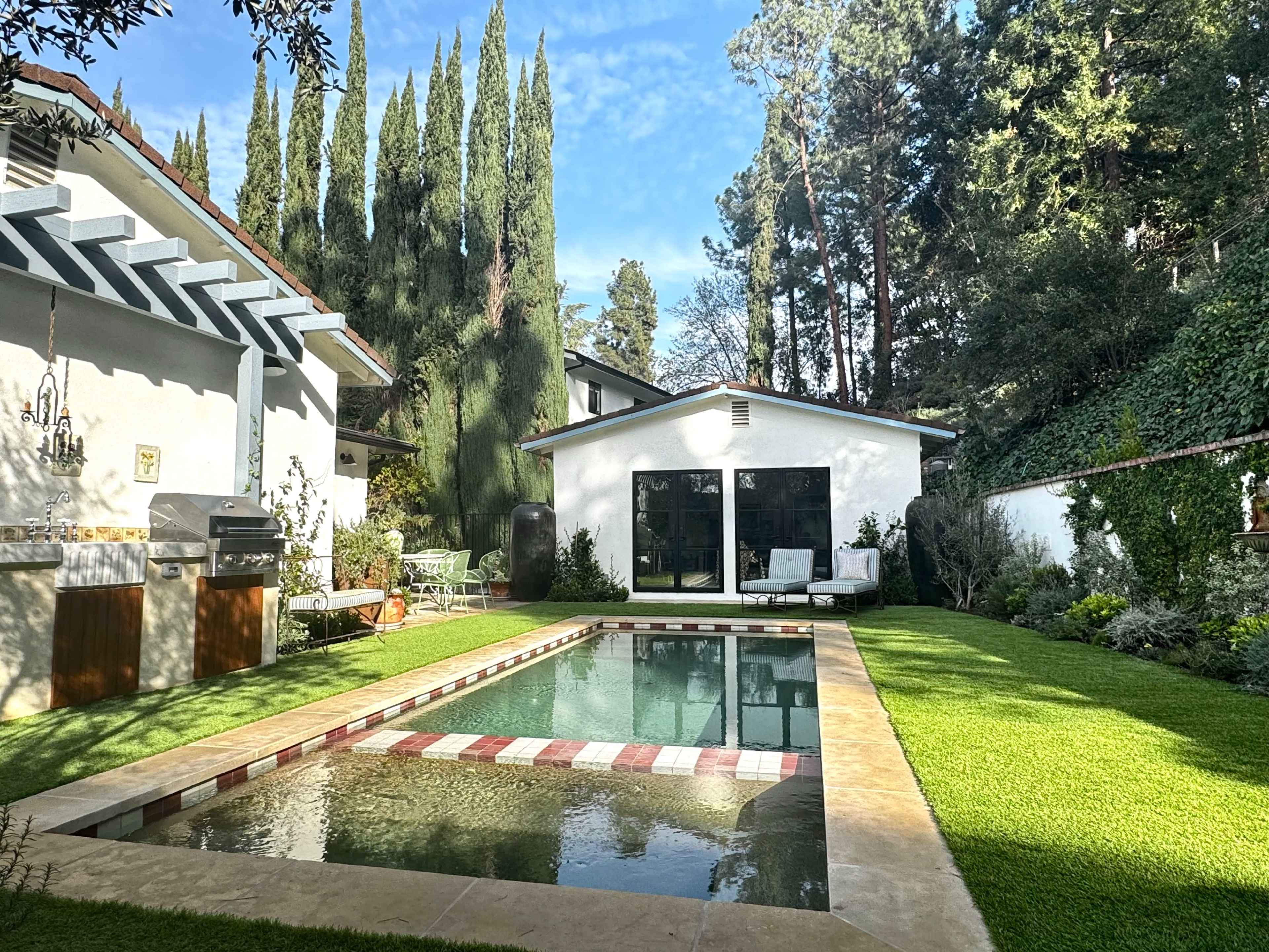 The image shows a backyard with a swimming pool surrounded by manicured grass and tall trees, with a modern home and outdoor seating area in the background.