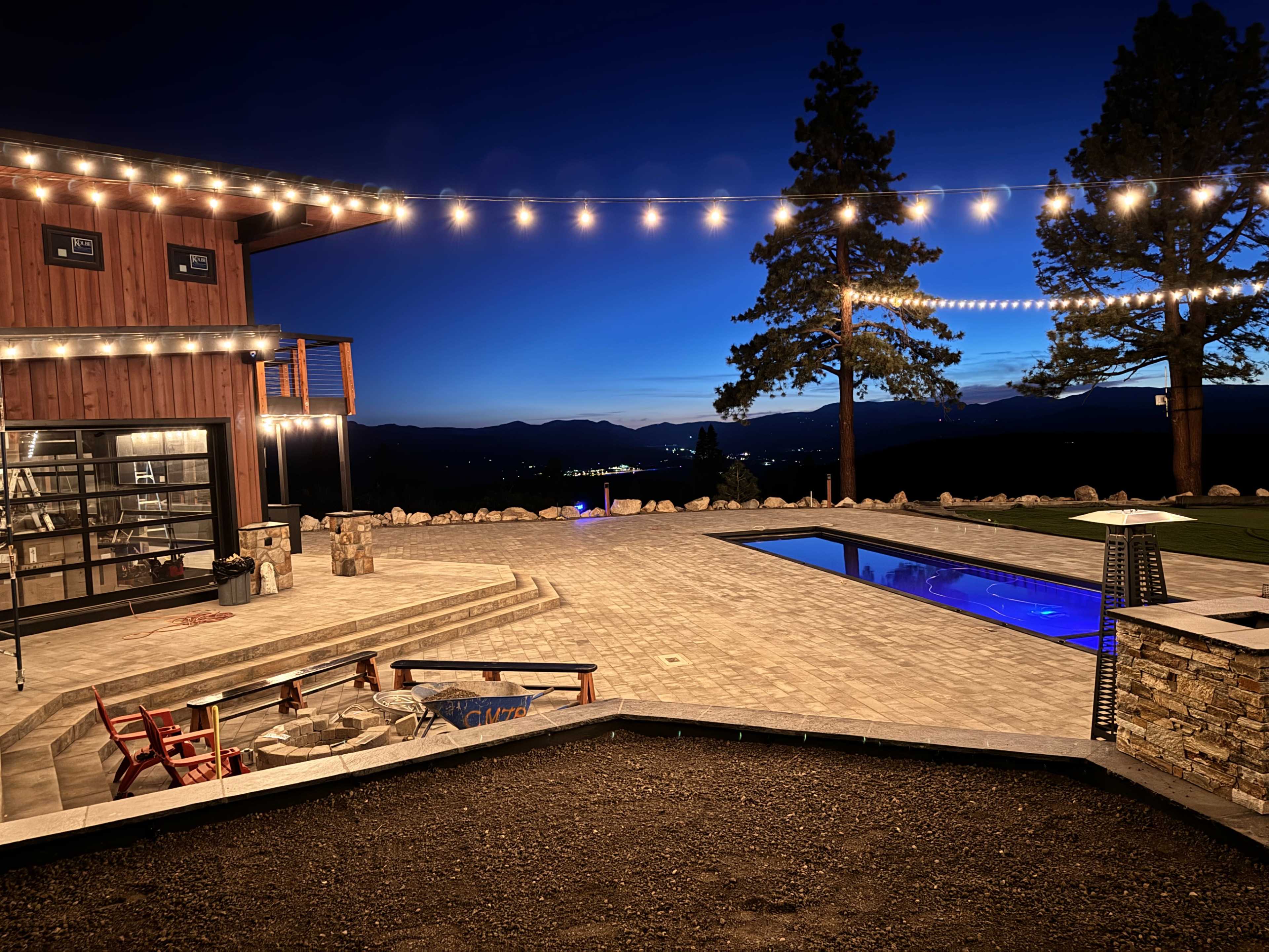 The image shows a modern outdoor patio with a pool, fire pit, and decorative string lights against a backdrop of trees and mountains at dusk.
