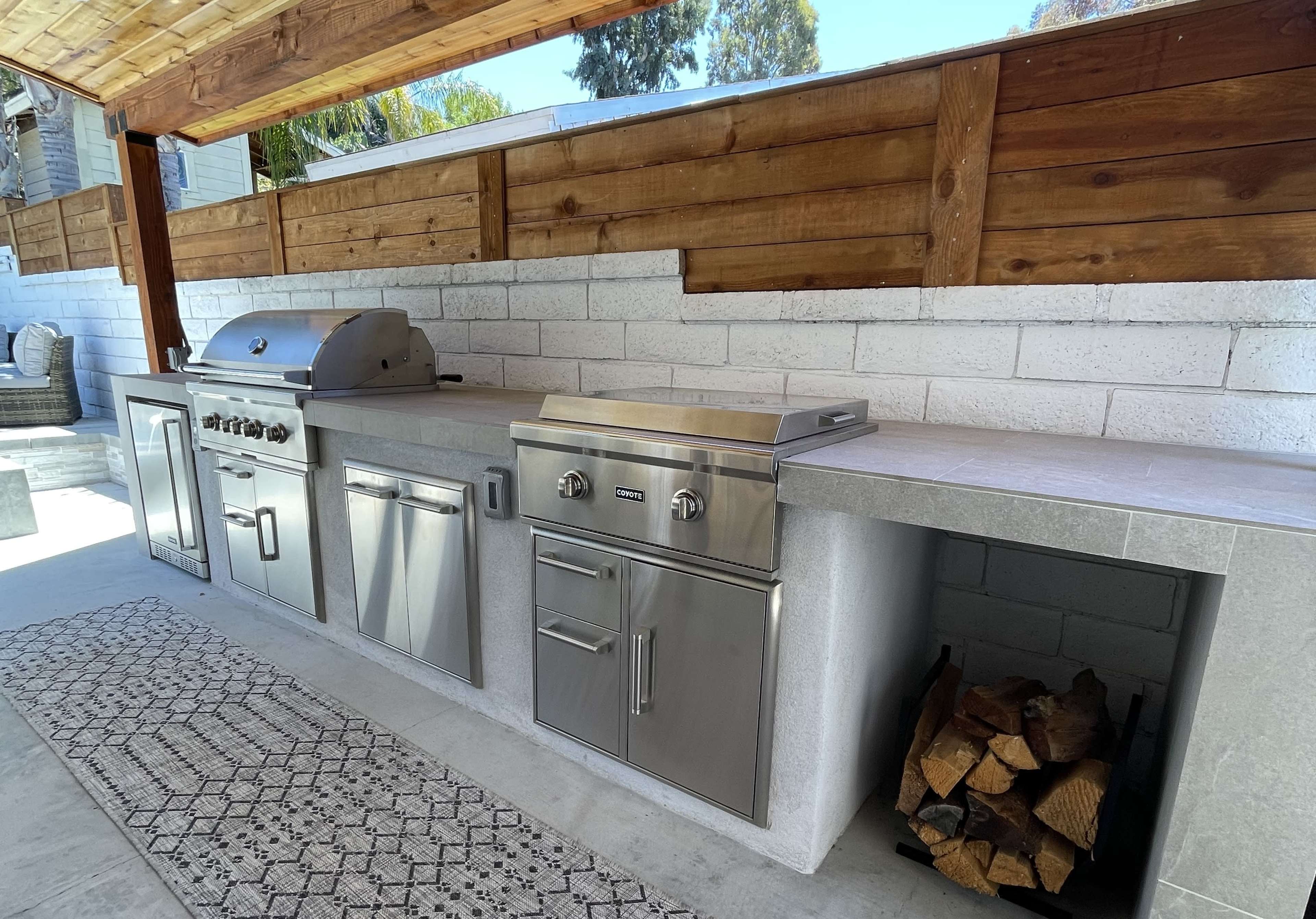 An outdoor kitchen featuring stainless steel appliances, including a grill and a countertop, against a backdrop of wooden panels and a stone floor.