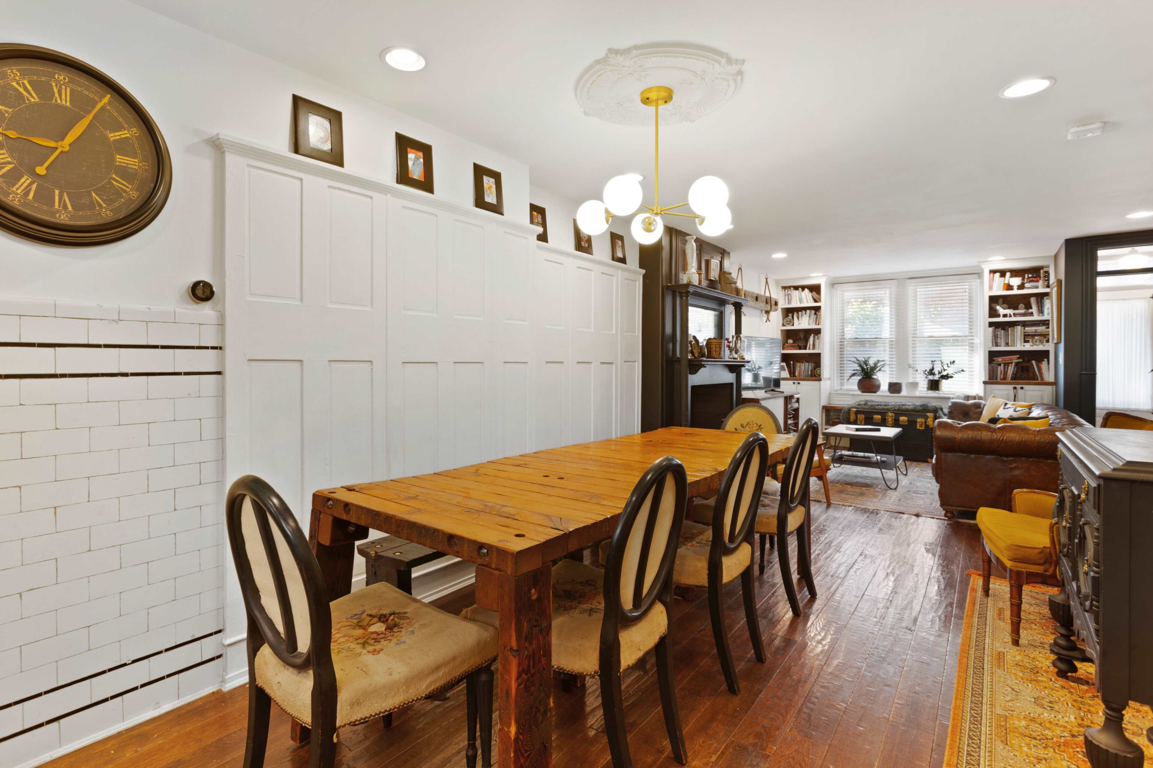 A dining area with a wooden table surrounded by six chairs, a large clock on the wall, and shelves filled with books in a well-lit room.