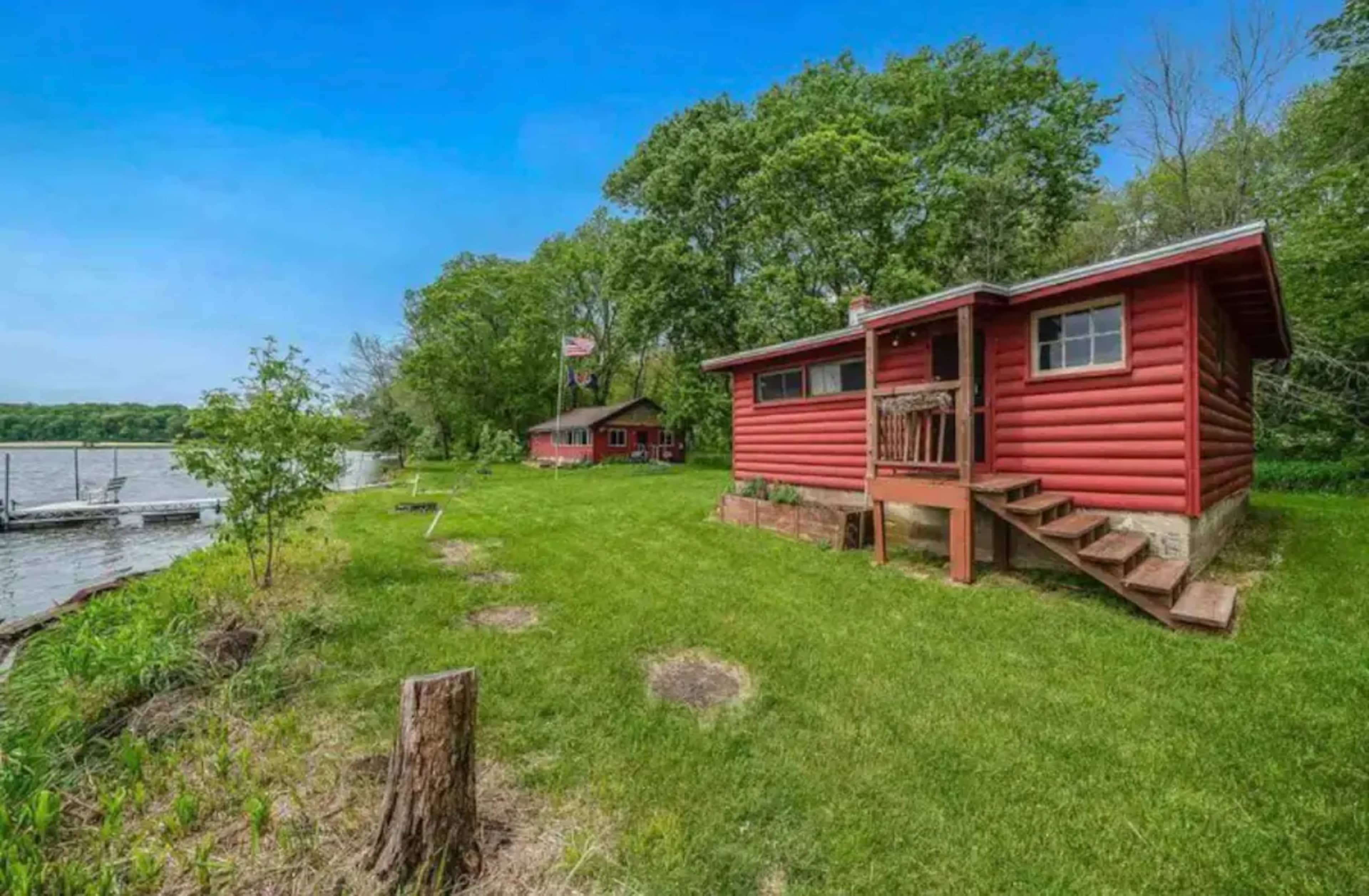 A red log cabin with a porch is situated near a lake, surrounded by trees and a grassy area.
