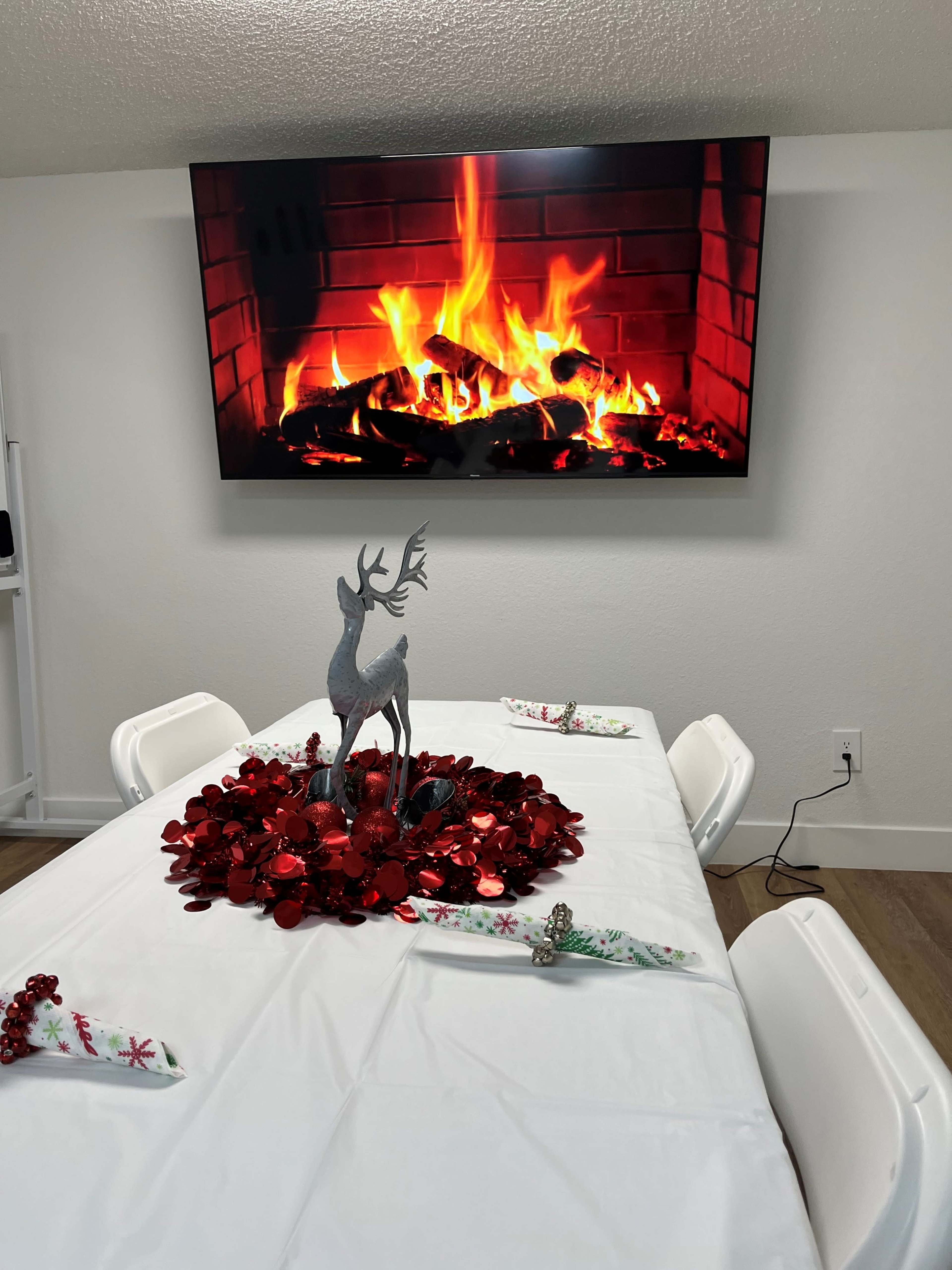 A dining table is set with a red and white themed centerpiece featuring a reindeer, with a large television displaying a fireplace above it.