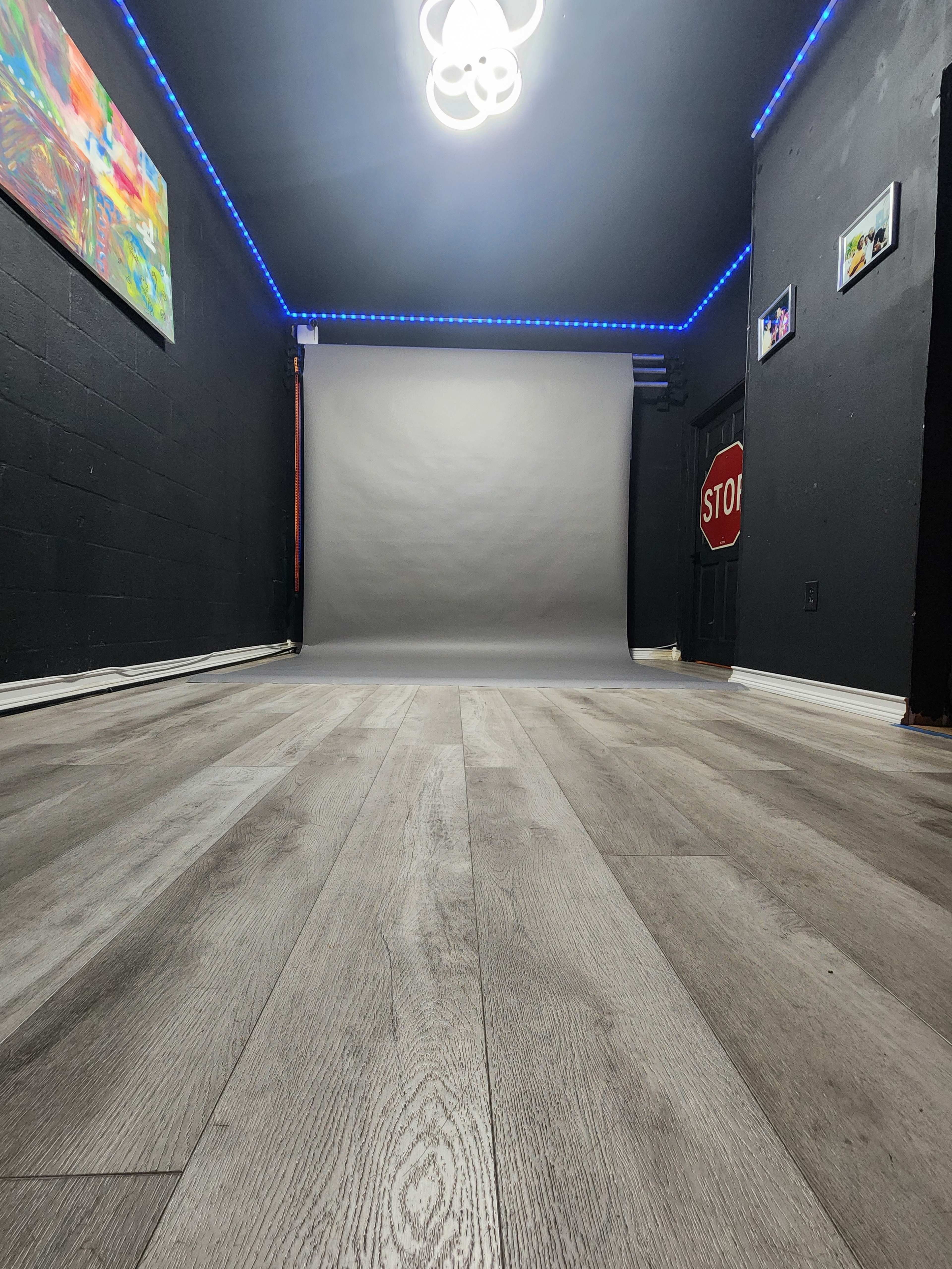 The image shows a spacious photo studio with gray flooring, black walls, and a lit backdrop framed by blue LED lights.