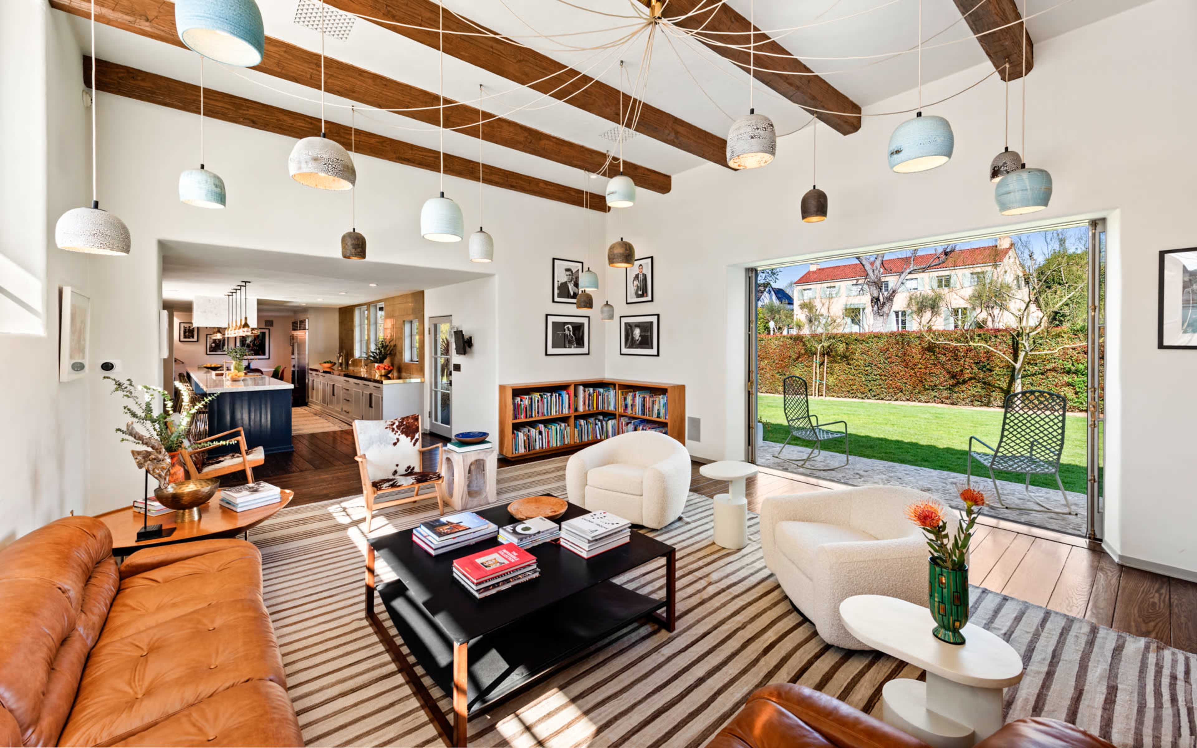 The image features a modern living room with brown leather sofas, a coffee table, bookshelves filled with books, and large windows that open to a green garden, all complemented by unique hanging light fixtures.