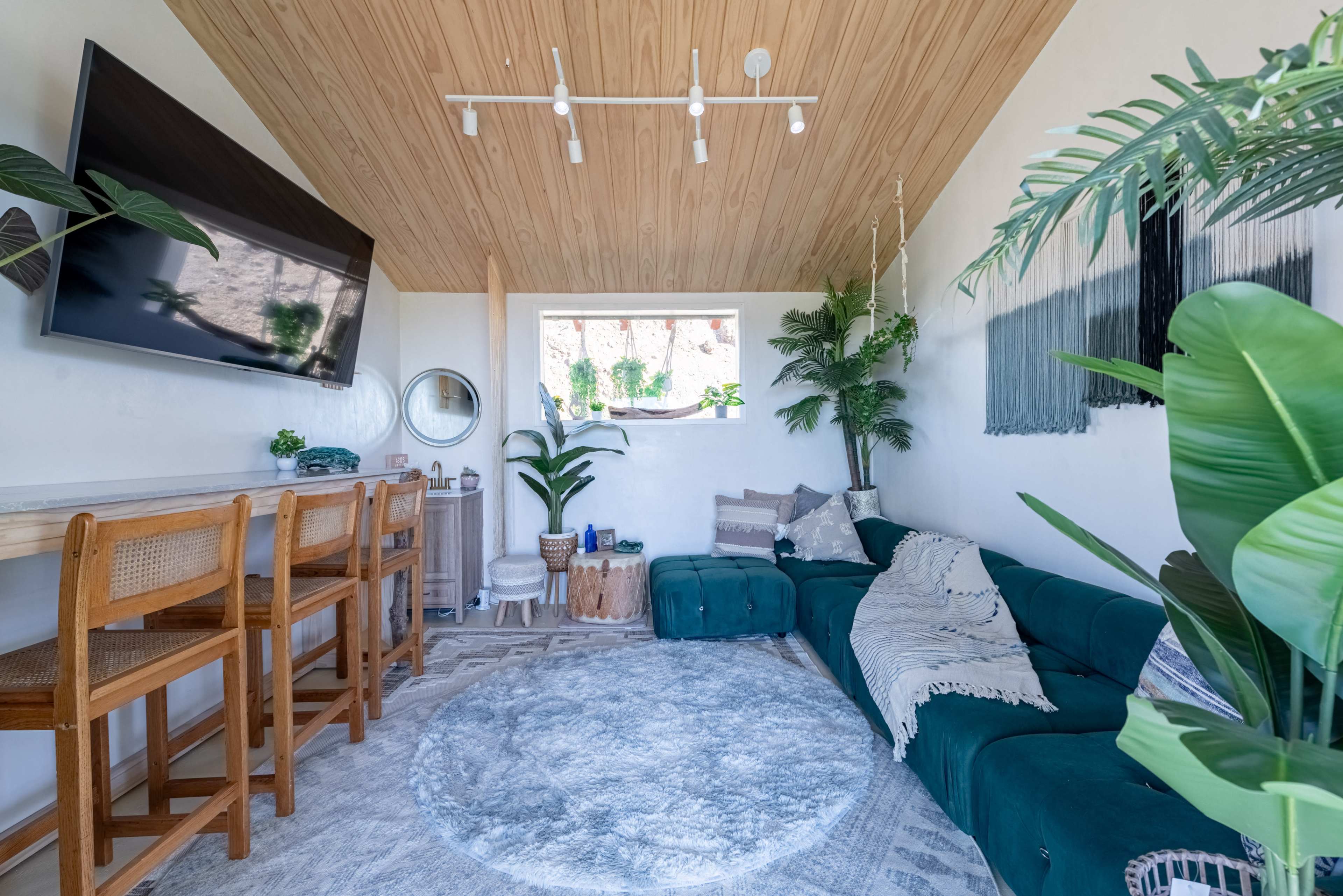 The scene features a modern living room with a green sectional sofa, a large round rug, a wooden bar with high stools, and plants decorating the space.