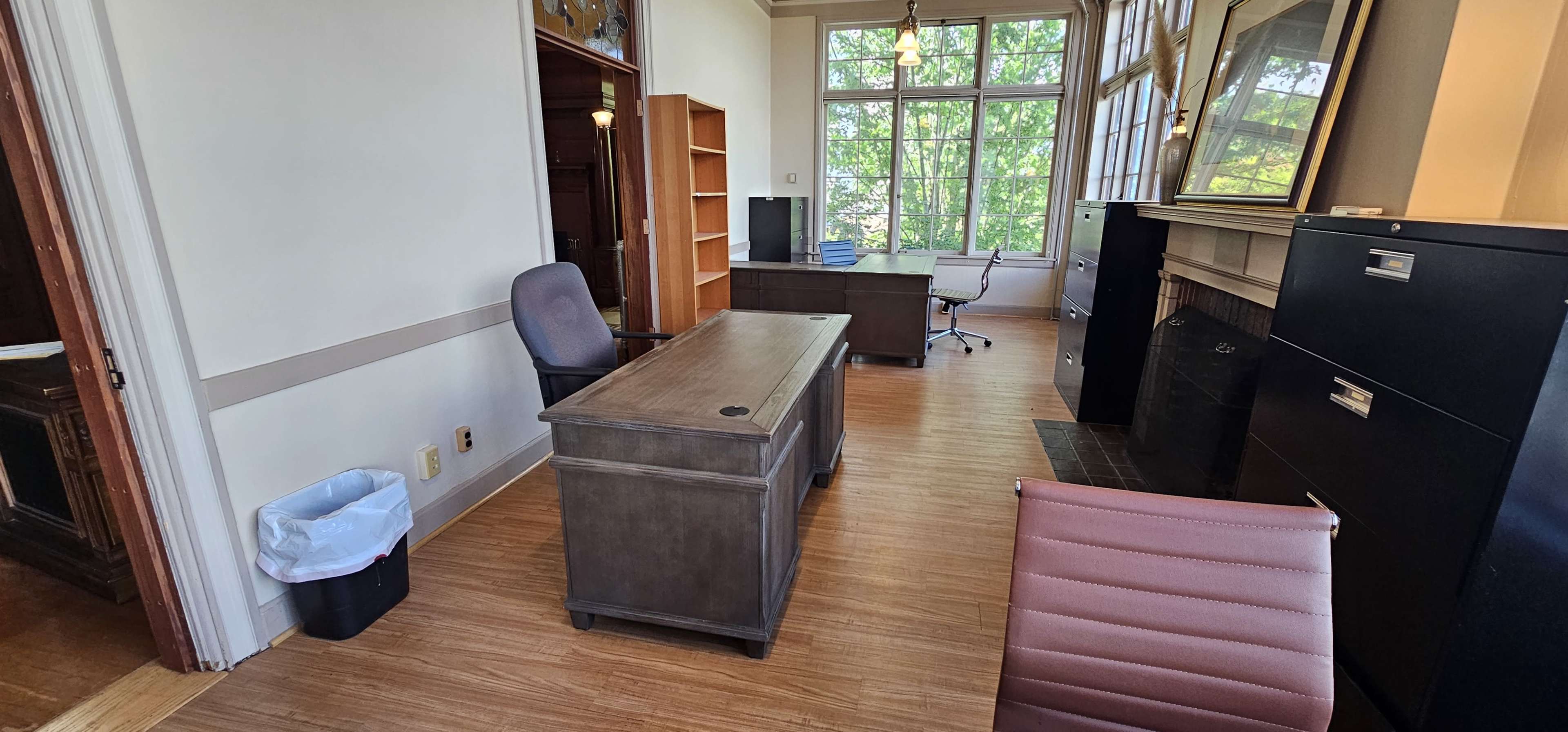 The image shows an office space featuring a wooden desk, a gray office chair, a black filing cabinet, bookshelves, and large windows with trees visible outside.