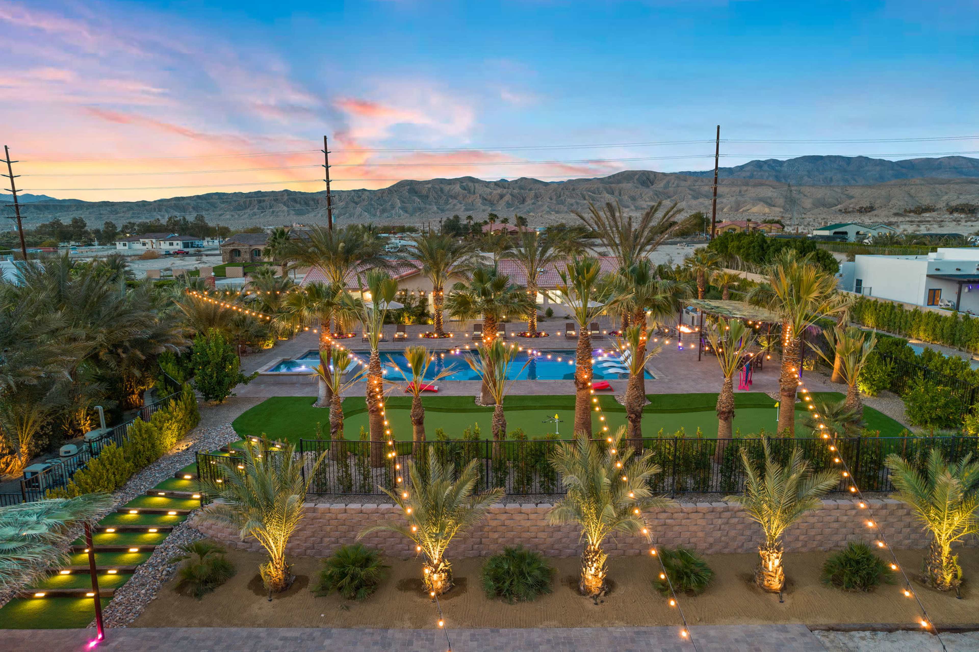 A landscaped outdoor area featuring palm trees, a swimming pool, and decorative lights against a backdrop of mountains and a colorful sky.