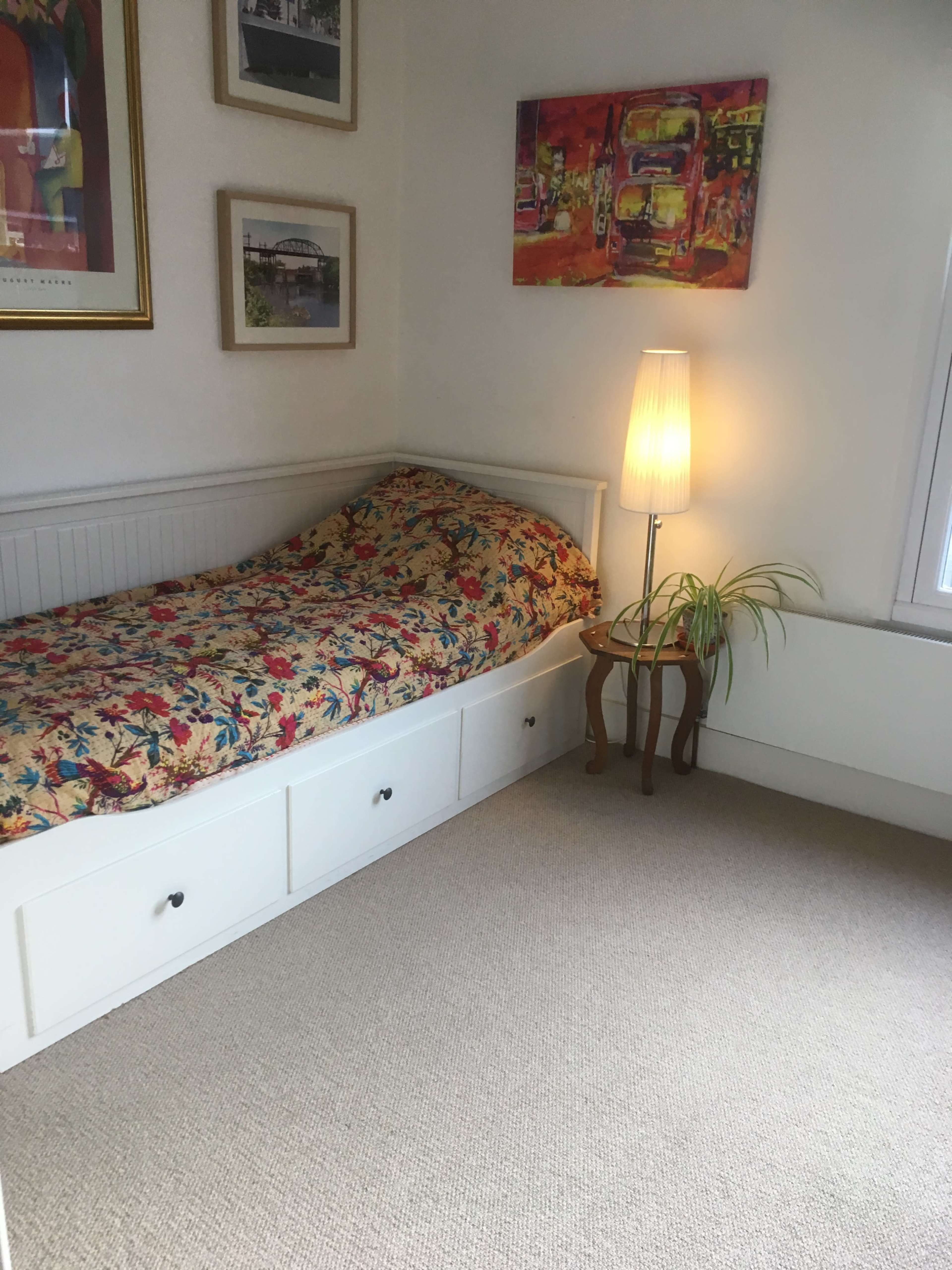 A small bedroom with a floral-patterned bedspread on a white bed, a standing lamp, and framed artwork on the walls.