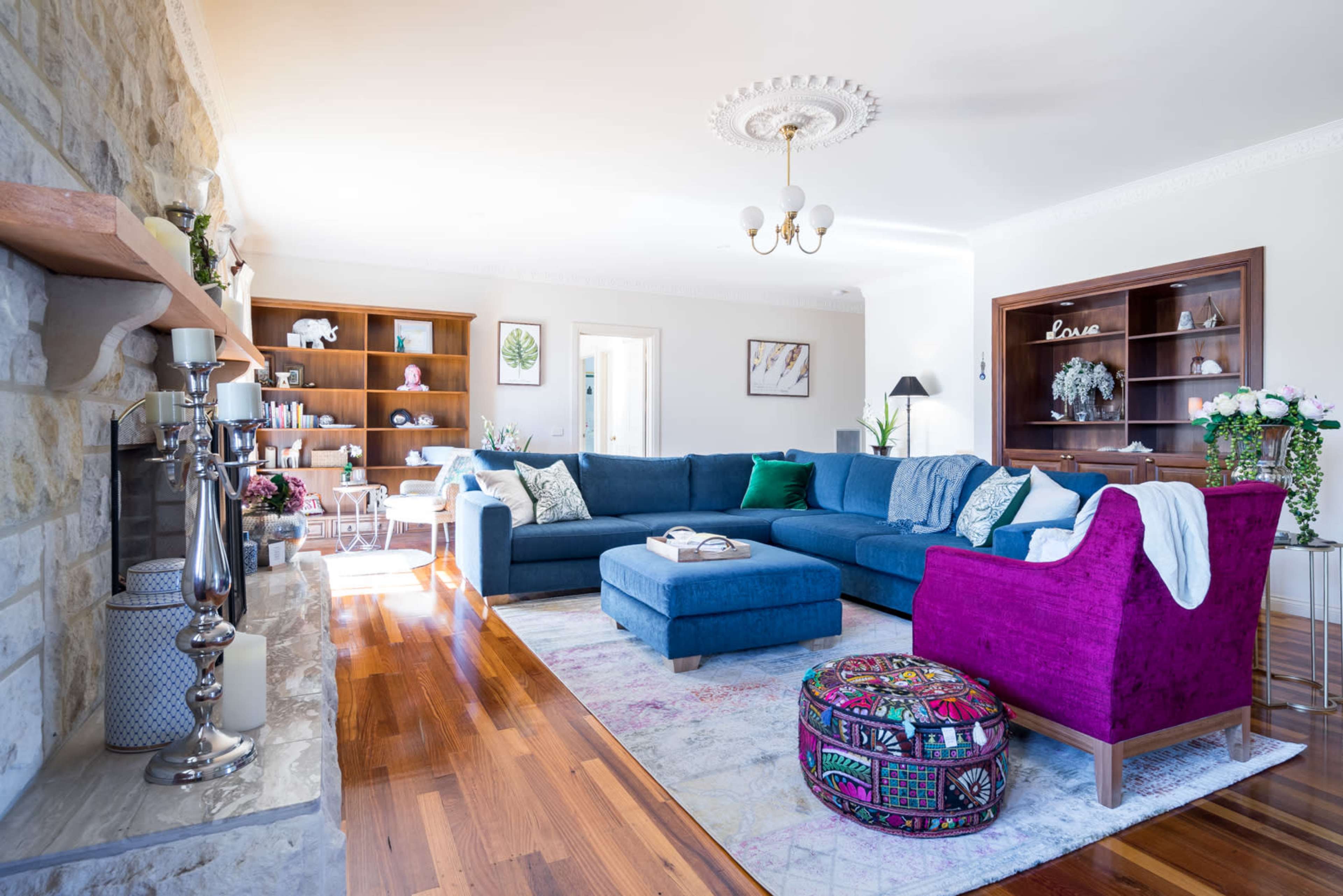A spacious living room features a large blue sectional sofa, a wooden coffee table, and colorful accent furniture, with natural light streaming through the windows.