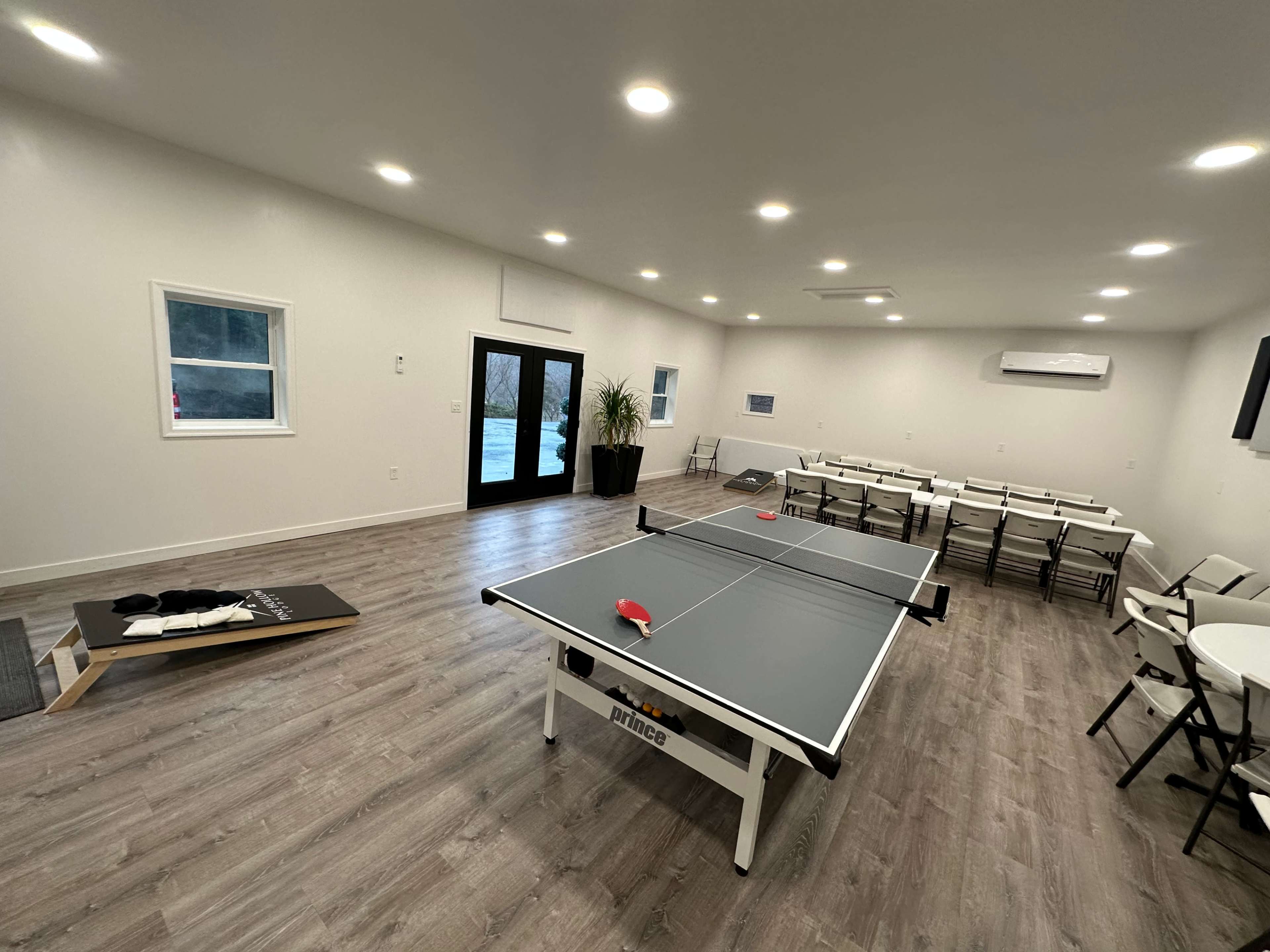 A spacious room features a ping pong table, seating arrangements with folding chairs, and a cornhole game setup in the corner.