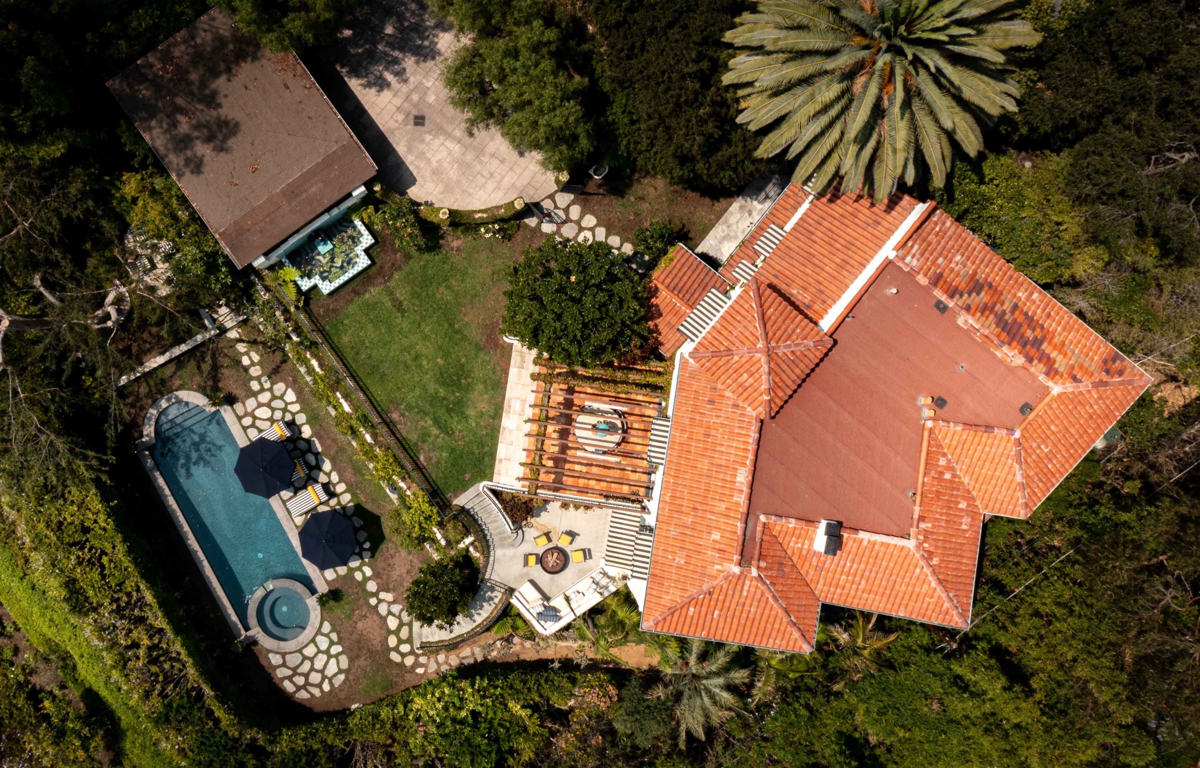An aerial view of a house with a red-tiled roof, surrounded by a landscaped garden, a swimming pool, and outdoor seating areas.