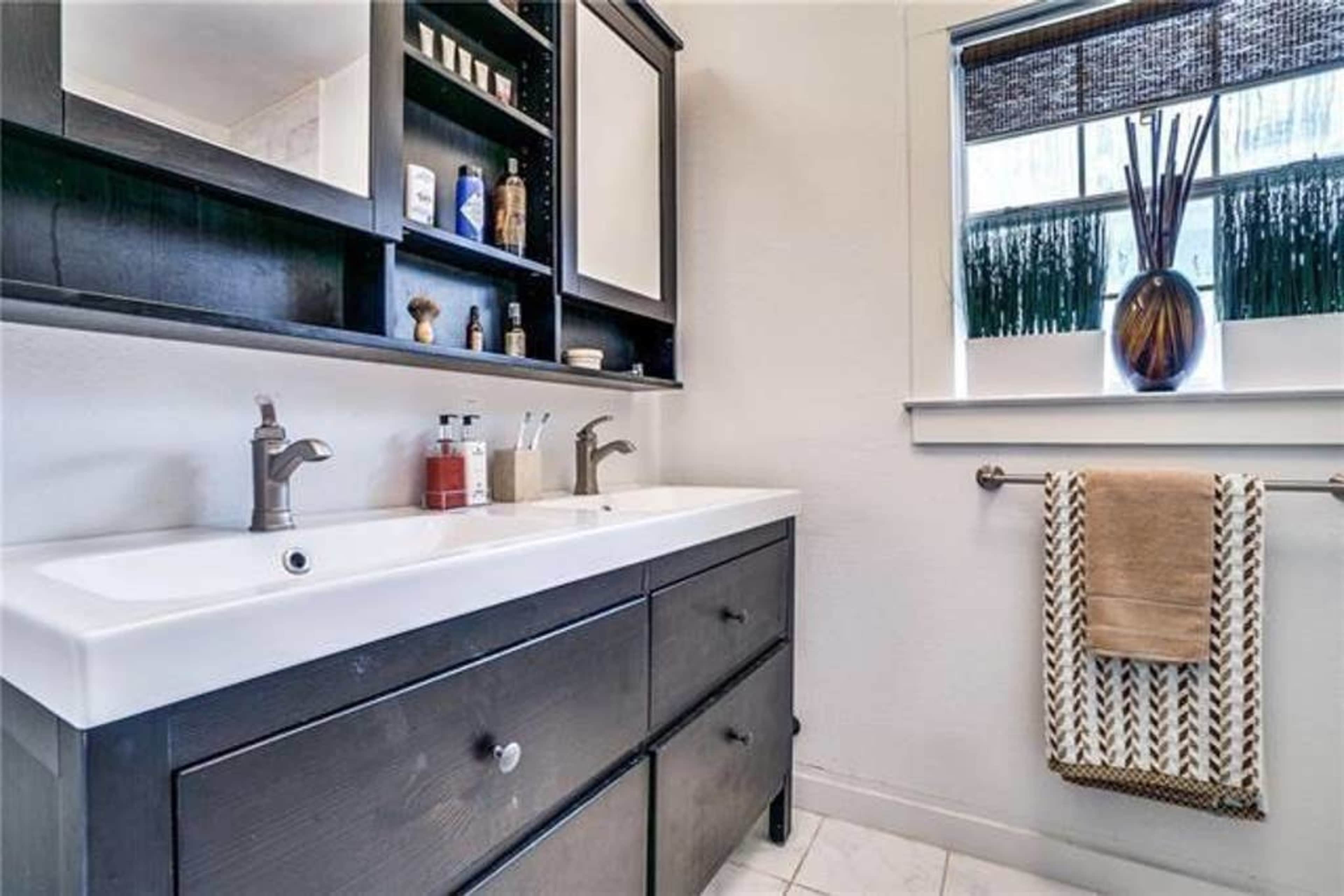 A modern bathroom with a dual sink vanity, dark cabinetry, and a window adorned with decorative plants.