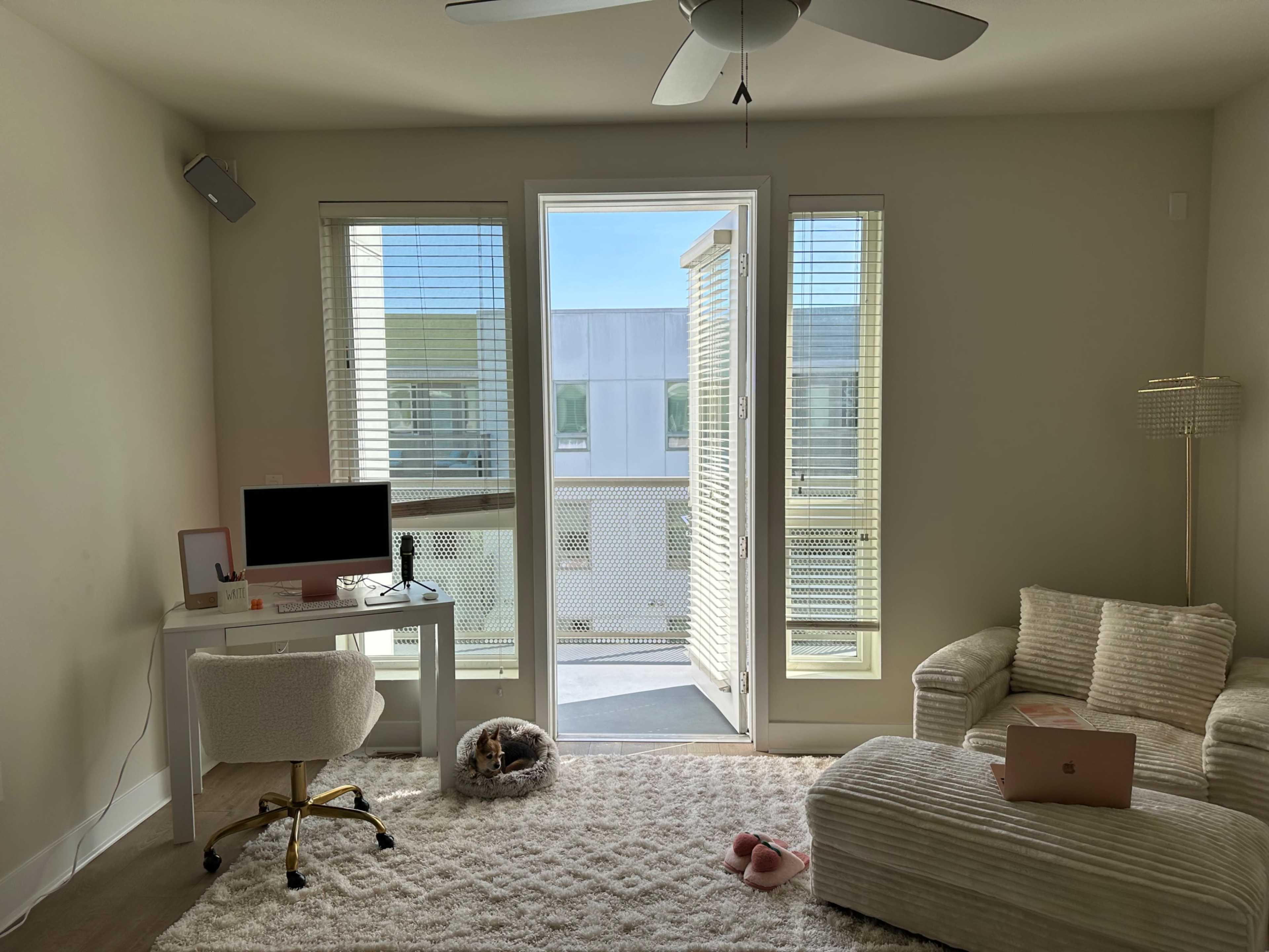 The image shows a light-colored room with a desk featuring a computer, a chair, and a cozy sofa, along with large windows leading to a balcony.
